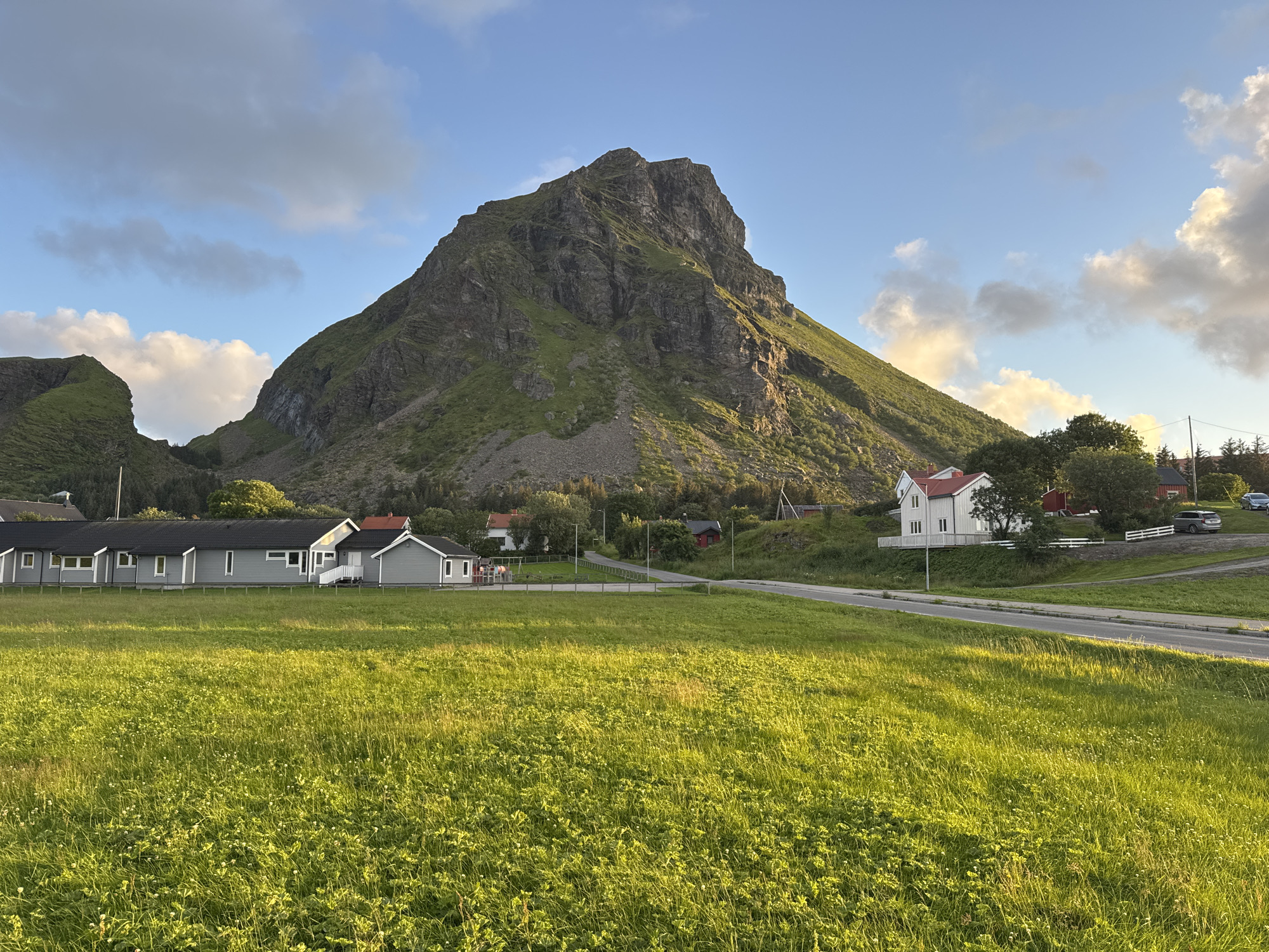 The image shows a rural landscape in Lovund, Norway, dominated by a large, rugged mountain with steep, rocky slopes and patches of green vegetation. In the foreground, a vast, lush green field stretches out, dotted with small white wildflowers. A paved road curves through the scene, separating the field from a cluster of houses.
To the left of the road, a row of modern, gray houses with dark roofs is visible. These appear to be residential or possibly holiday homes, with neat, manicured lawns in front. Further to the right, a larger white house with a red roof stands on a slightly elevated plot, bordered by a white fence. Beyond this, other buildings and trees are scattered across the landscape, leading towards the base of the mountain.
The sky is a clear blue with scattered white and gray clouds, suggesting fair weather. The sunlight casts long shadows and highlights the textures of the mountain and the field, indicating it is likely late afternoon or early evening. The overall impression is one of serene, natural beauty in a remote, mountainous coastal region. There are no people visible in the image.