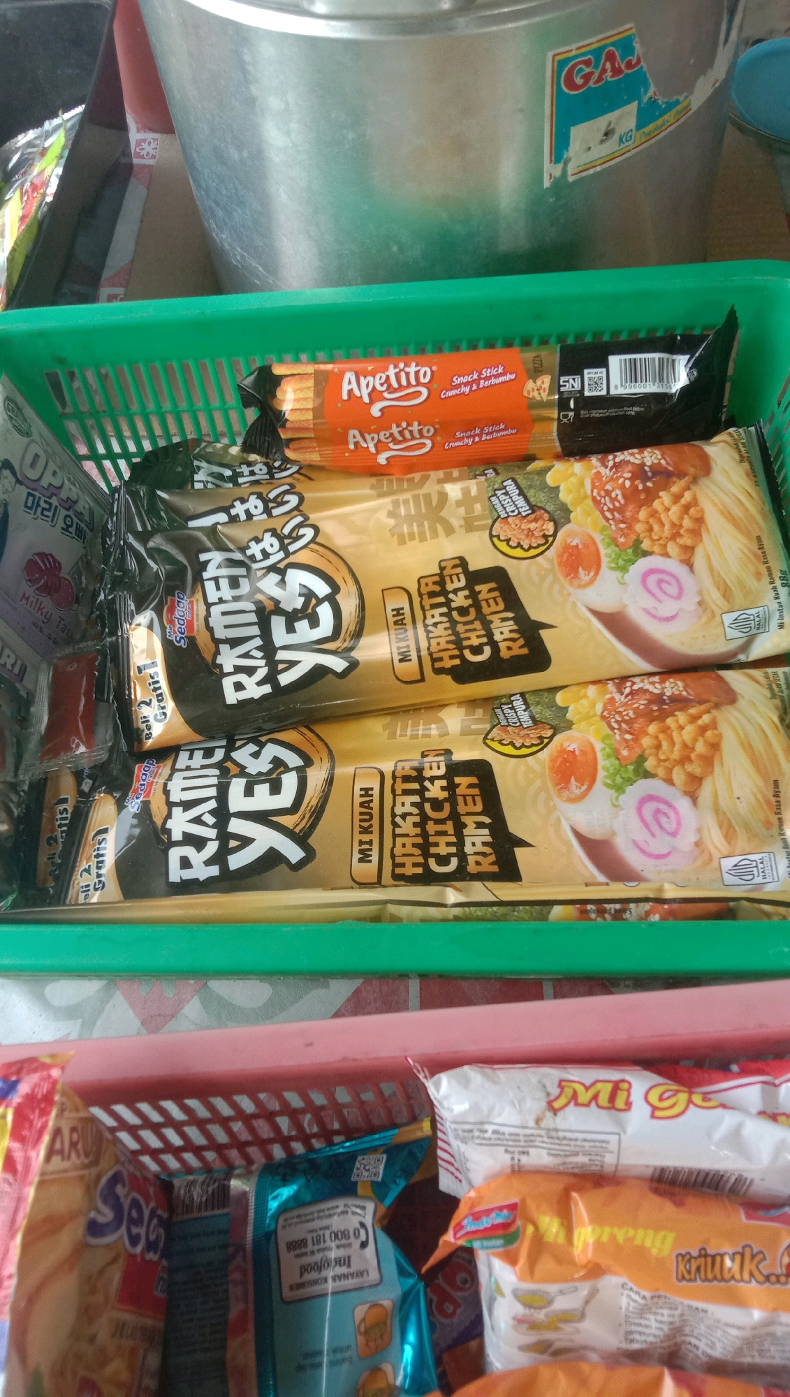 The image shows a top-down view of two green and pink plastic baskets filled with various food items. The green basket, in the foreground, contains several packages of instant noodles and snacks. Most prominent are two identical packages of "Ramen Yes" with a Hakata Chicken flavor. Above these, a package of "Apetito" snack sticks is visible. To the left of the ramen packages, other smaller packages of snacks and what appear to be condiment sachets are present, some with Korean text. The pink basket, positioned behind the green one, holds more food items, including bags of "Mi G" instant noodles and other packaged snacks. In the background, a large metallic container or pot is partially visible, with a sticker on its side featuring the text "GAJAH" and "KG". The overall setting appears to be an indoor environment, possibly a pantry or storage area, with a tiled surface visible beneath the baskets. The image is taken from a high angle, looking down into the baskets. There are no people visible in the image. The location is identified as Ngempit, Indonesia.