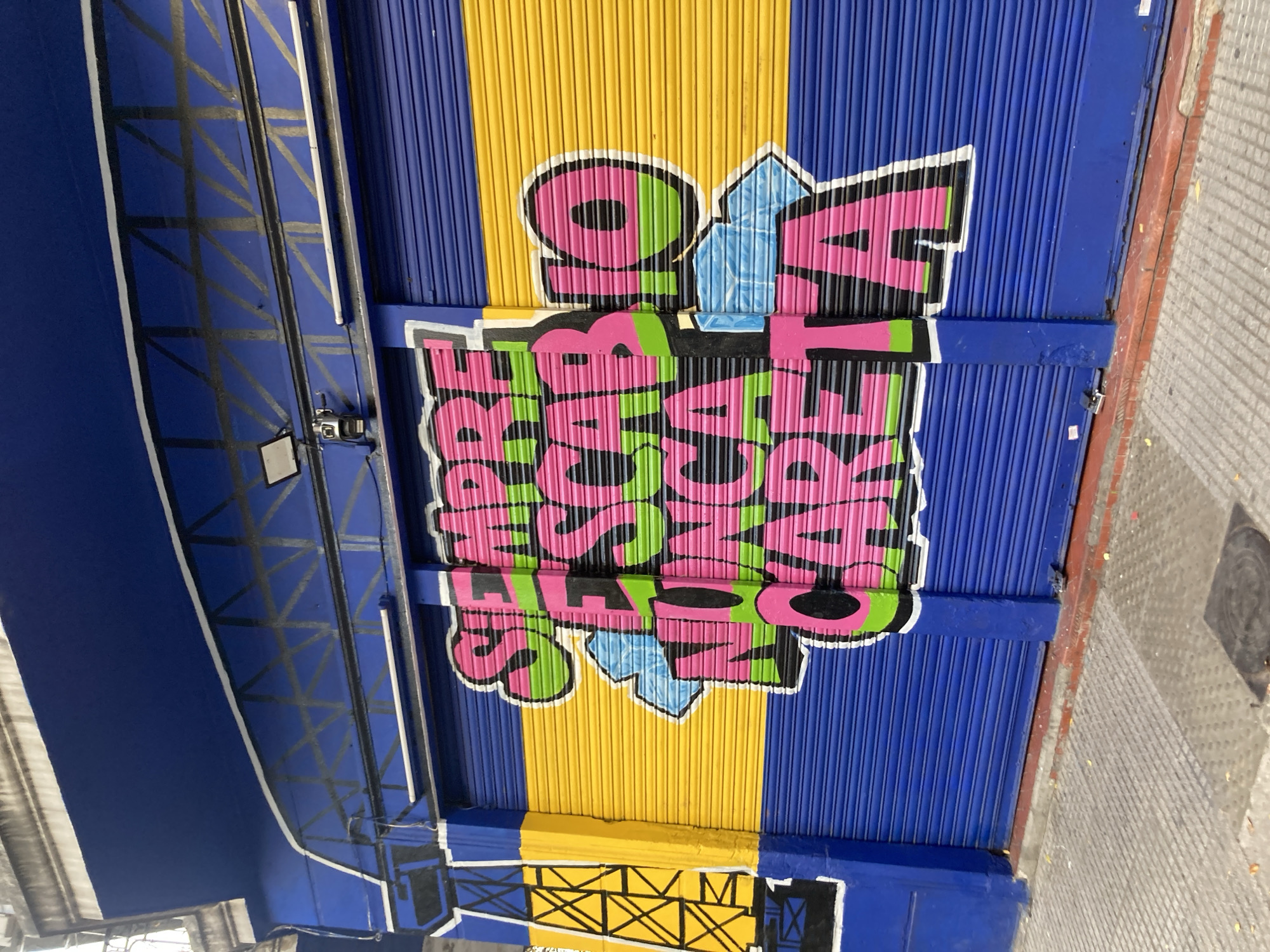 The image shows a colorful mural on a corrugated metal roll-up door or wall. The background is striped horizontally in yellow and blue. The mural itself features the words "SUPER SABADO DE NOTICIAS" stacked vertically and in bright pink and green colors with a white outline. Small blue, diamond-shaped elements separate the words. The roll-up door is located in Buenos Aires, Argentina. The building has a blue metal framework above the wall and a gray sidewalk is visible on the right side of the frame.