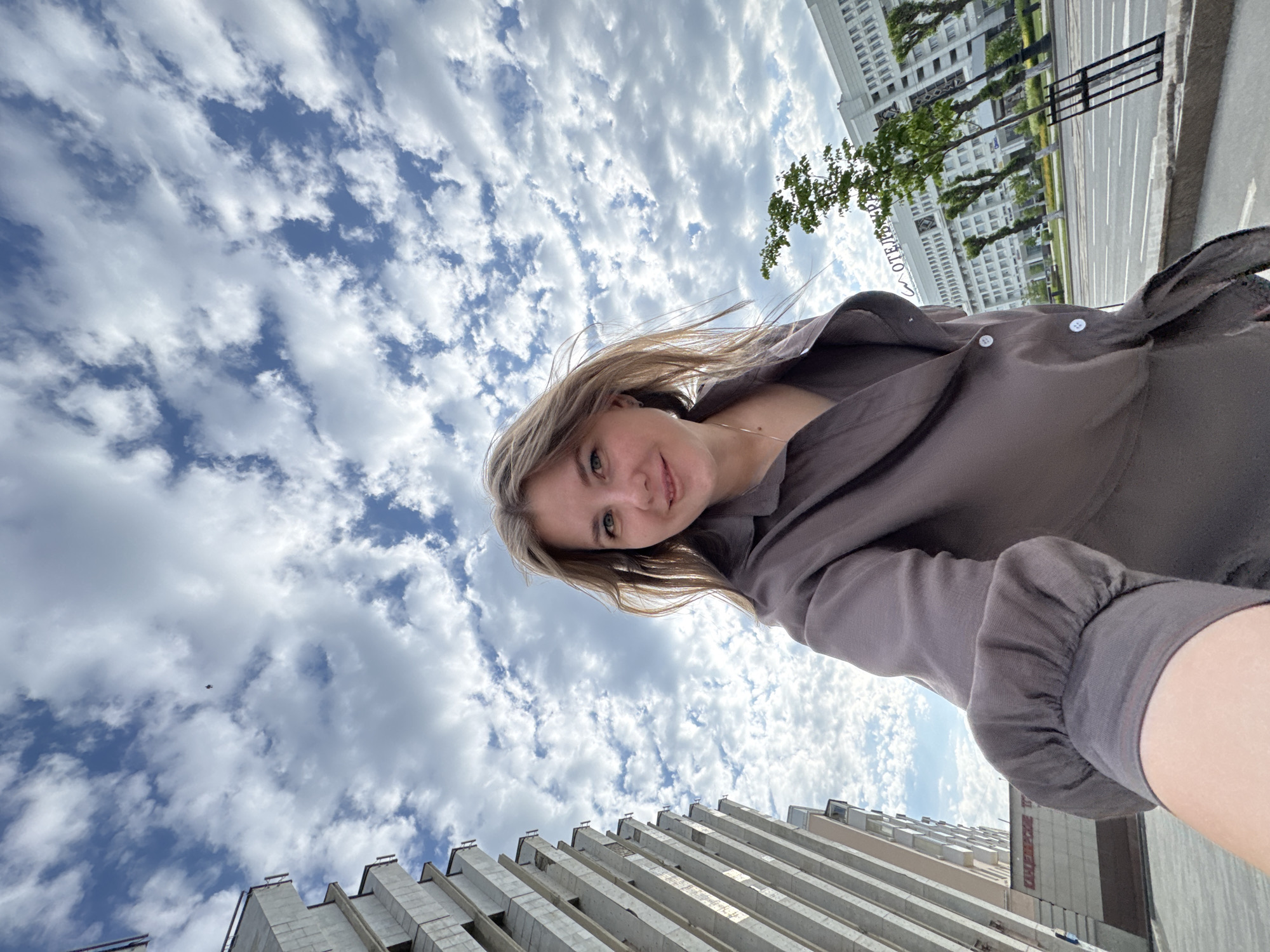 A selfie-style photograph shows a fair-skinned woman with light brown hair and green eyes wearing a brown button-up shirt. The shot is taken from a low angle, looking up at the woman and the sky, which is filled with cumulus clouds. In the background, the tops of buildings are visible. A building has the Cyrillic word "OTEL" visible. This places the location as Perm, Russia, specifically near the Perm Hotel.