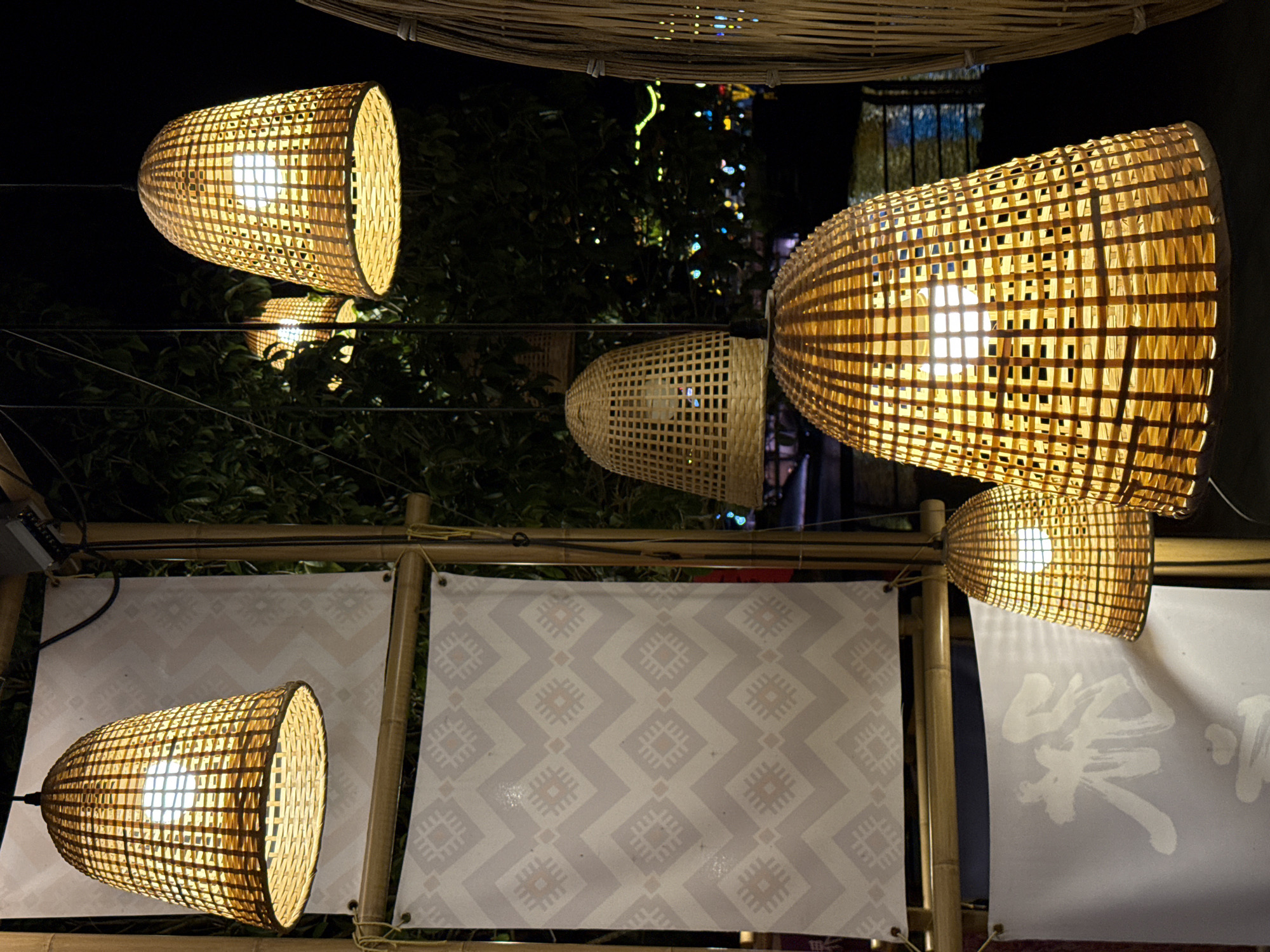 The image presents an illuminated outdoor night scene in Wulingyuan, China, showcasing a vibrant display of traditional bamboo lanterns and decorative banners. The central subjects are numerous handcrafted lanterns made from intricately woven bamboo strips, glowing brightly with warm yellow light. These lanterns vary in shape, including elongated cylindrical and more rounded or conical forms, suspended at different heights and angles, dominating the mid-ground and foreground.
A sturdy framework of bamboo poles and horizontal bars supports both the lanterns and several rectangular banners below them. Two prominent banners are visible: one features a repeating light gray and white geometric pattern, possibly a traditional textile design. Adjacent to it, another banner displays elegant white Chinese calligraphy against its light background, although the specific characters are not fully legible.
In the background, dark green foliage from trees is subtly illuminated by the lanterns, with hints of bokeh lights suggesting distant festive or urban lighting. A large, flat bamboo woven structure is visible at the very top of the frame, possibly part of a ceiling or canopy.
The time of day is clearly night, as indicated by the dark background and the reliance on artificial light from the lanterns. The overall atmosphere is warm, inviting, and festive, suggesting a market, restaurant, or cultural display. No people are visible, and no specific activity is captured beyond the display itself. The traditional bamboo craftsmanship and calligraphy provide strong cultural cues, consistent with the specified location of Wulingyuan, China.