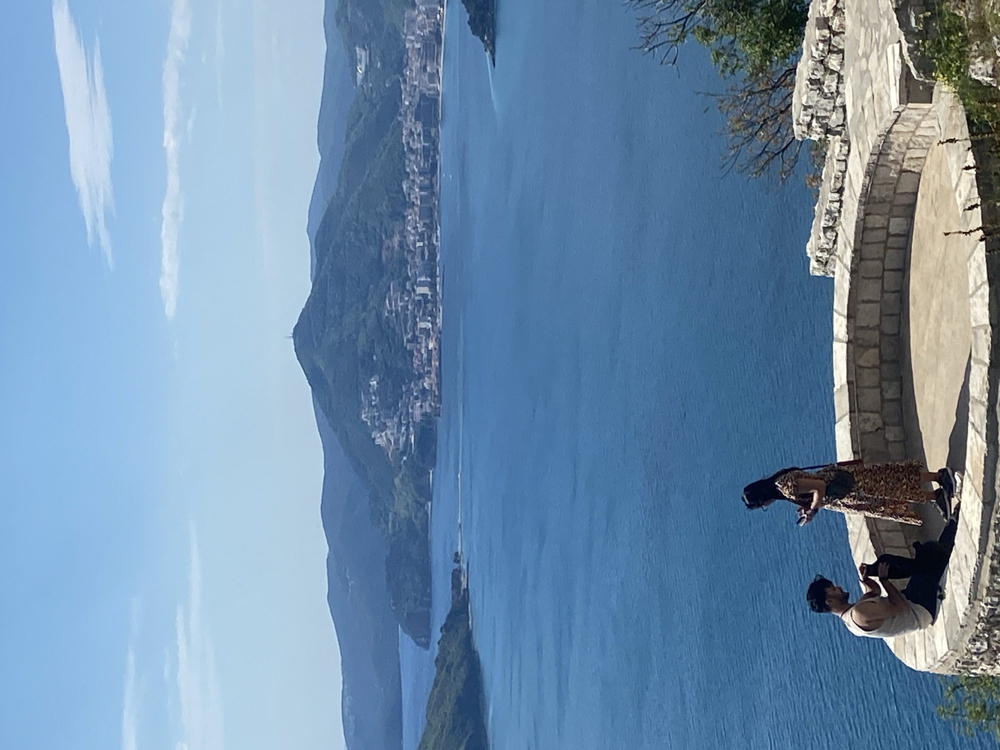 The image shows a view from a stone balcony overlooking the Adriatic Sea, likely in or near Sveti Stefan, Montenegro. Visible in the background are mountains and a portion of a coastal town. Two people are on the balcony. One person is sitting on the balcony taking a photo. The second person is standing and looking at the view. The sky is blue with some clouds.