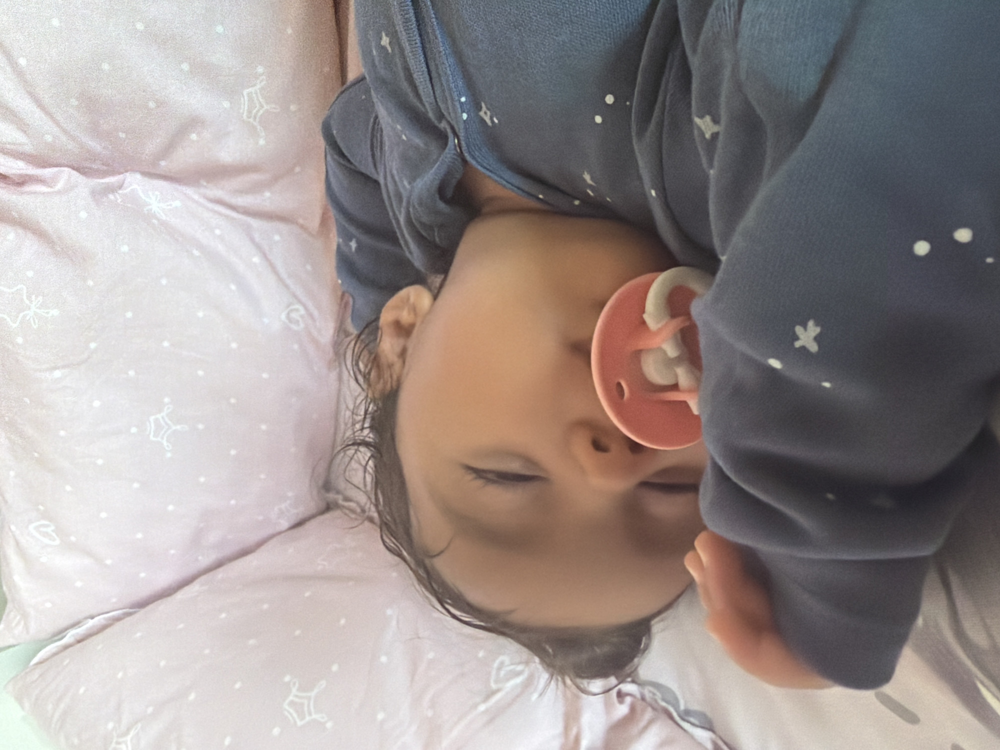 The image shows a close-up of a baby sleeping on a pink pillow with a white star pattern. The baby is wearing a dark blue onesie with white star and dot patterns. A pink pacifier is in the baby's mouth. The baby's eyes are closed, suggesting they are asleep. The lighting is soft, and the background is blurry, creating a peaceful and intimate atmosphere. The context provided indicates the location is Razhevo Konare, Bulgaria, but there are no visible cues in the image to confirm this. The image appears to be taken indoors, likely in a bedroom or nursery.