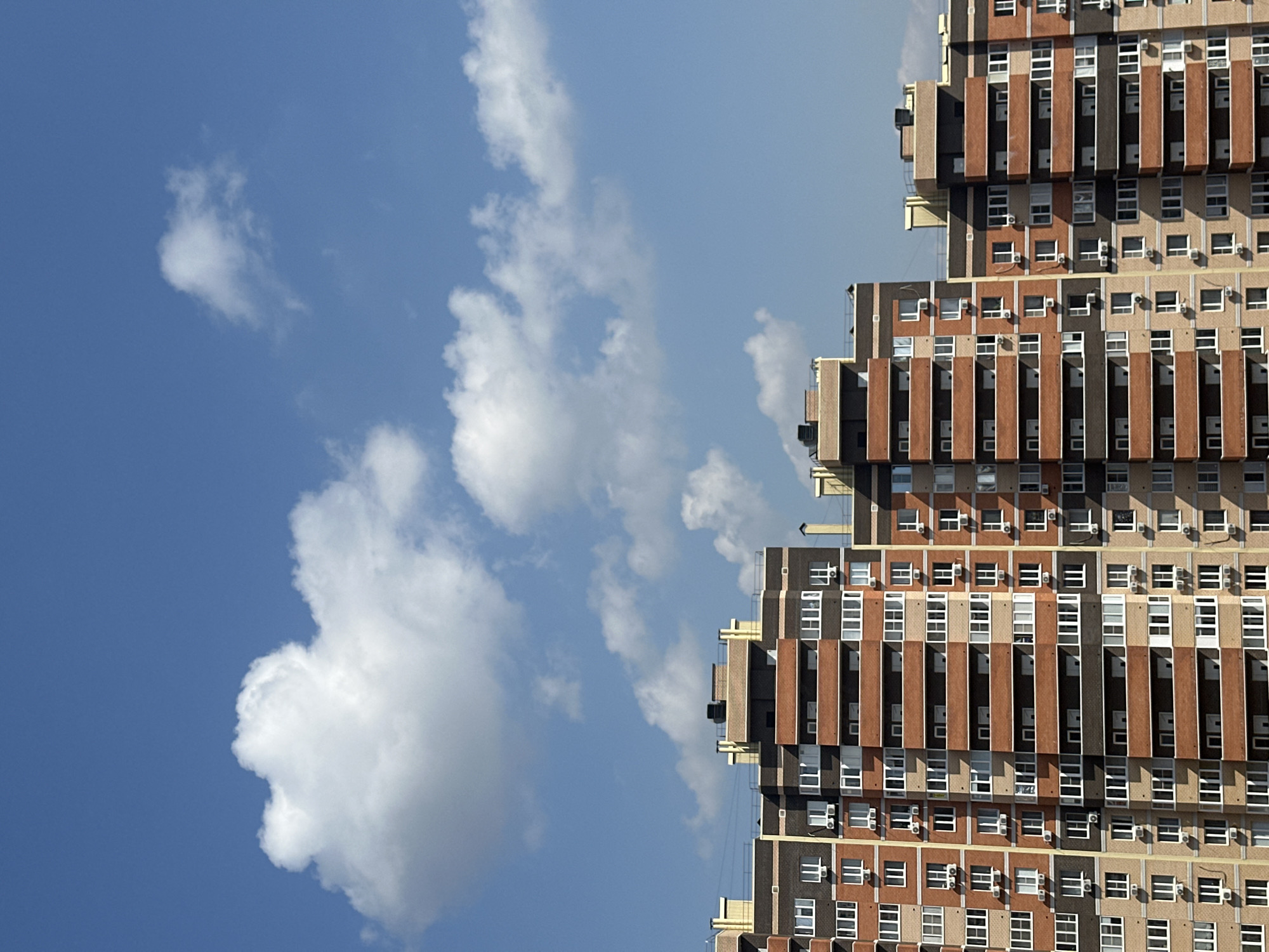 The image shows a high-rise apartment building against a blue sky with scattered clouds. The building is multi-story with a repetitive pattern of windows and balconies. There are air conditioning units visible on many of the balconies. The building has a layered or stepped design, with each successive floor set back slightly from the one below. The colors of the building are primarily brick red, brown, and concrete gray. The sky is a light blue color with white, fluffy clouds. There is no indication of the location being Novoaleksandrovka, Russia.