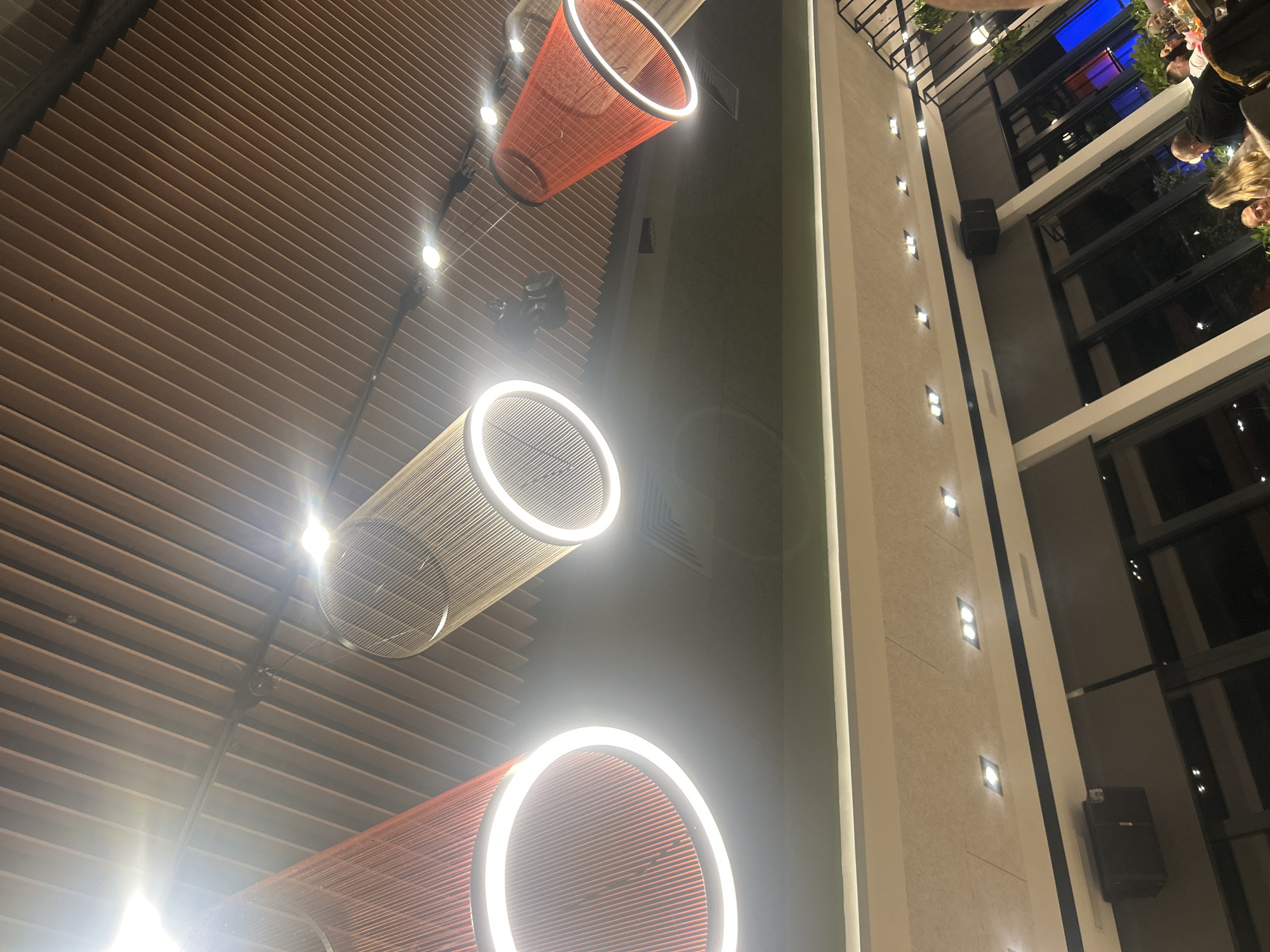 The image shows an interior space with distinctive design elements. A horizontally striped ceiling is visible, punctuated by modern, cylindrical light fixtures, some of which are white and others red. The lights are a prominent feature, with a few strung along the ceiling. To the right of the image, there's a high wall with evenly spaced, small square lights running vertically along its surface. The wall is part of a structure with large windows, suggesting a multi-story building. Through the windows, there are hints of an outdoor setting with foliage and what appears to be a restaurant or gathering space with people present, possibly dining. The scene is indoors with a modern design aesthetic. The specific location in Plovdiv, Bulgaria, cannot be determined from the image.