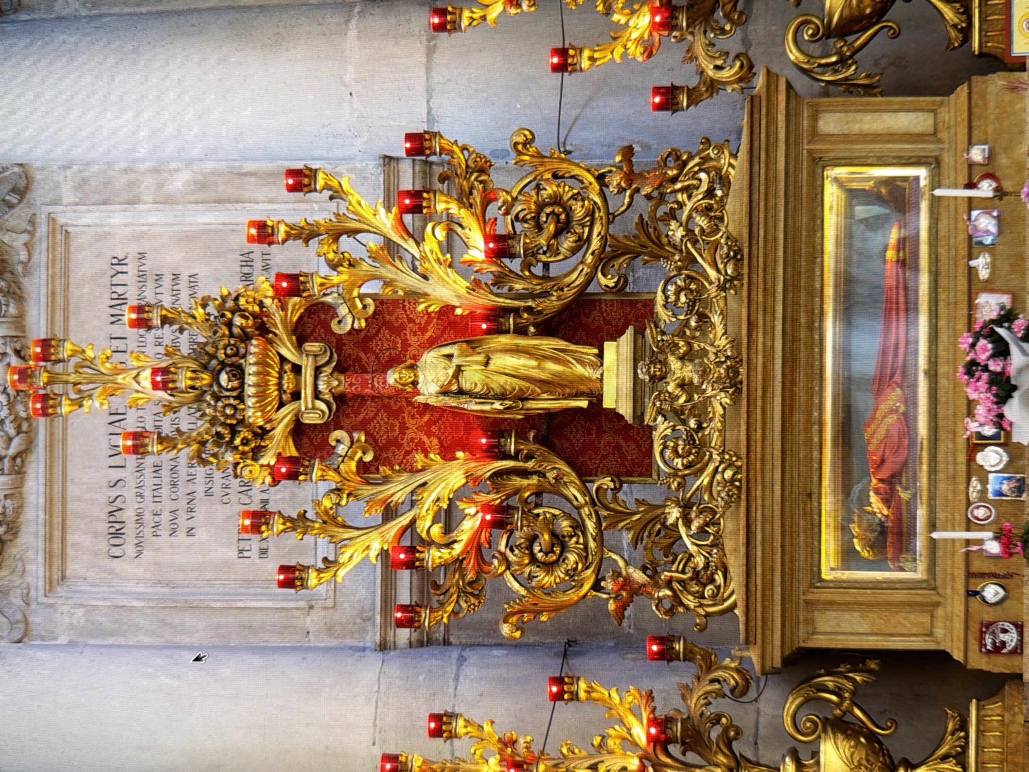 This is a full-frame, eye-level, medium shot of a golden altar and reliquary containing a mummified body. The reliquary is made of wood with a glass display case. The body is dressed in red and gold robes and appears to be a female saint. Above the reliquary, a golden altar decoration features multiple red candles, as well as a statue of a golden robed figure, likely Saint Lucy, who is the patron saint of the blind. The inscription on the wall behind the altar reads "CORPUS S. LUCIAE ET MARTYR". The scene is lit by ambient light and the glow of the red candles. This shrine is located in the Saint Nicholas Roman Catholic Cathedral in Kyiv, Ukraine.