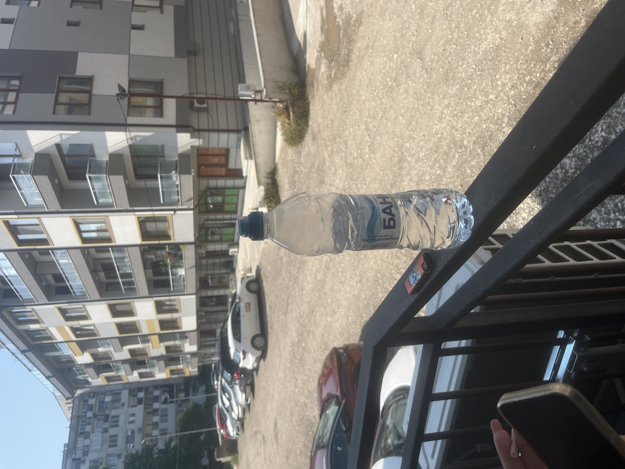 This image, taken in Varna, Bulgaria, captures a bright, sunny urban scene from an elevated vantage point, likely a balcony or terrace. In the immediate foreground, a clear plastic water bottle lies horizontally on a dark metal railing. The bottle's label, written in Cyrillic, prominently displays "БАНЯ" (Banya) and below it, "РЕЦИКЛИРАЙ МЕ" (Recycle Me). To the right, a hand with light-colored, manicured nails holds a gold-colored smartphone, partially visible behind the railing. A small, colorful item, possibly a decorative lighter or key fob, rests on the railing next to the bottle.
Beyond the railing, a parking area with an asphalt or light-colored paved surface is visible. Several cars are parked, including a white SUV-like vehicle, a red sedan, and other darker vehicles. Sparse patches of green vegetation grow along the edges of the parking area, and the ground appears dry and sun-drenched.
In the background, multi-story residential buildings rise under a clear blue sky. One building features a modern, modular design with varying shades of grey and white blocks, numerous windows, and glass-fronted balconies. Further in the distance, another, older-looking residential complex with many windows is also visible. The strong sunlight indicates it is likely midday or early afternoon, casting distinct shadows across the scene. The overall impression is that of a typical urban setting on a warm, clear day.