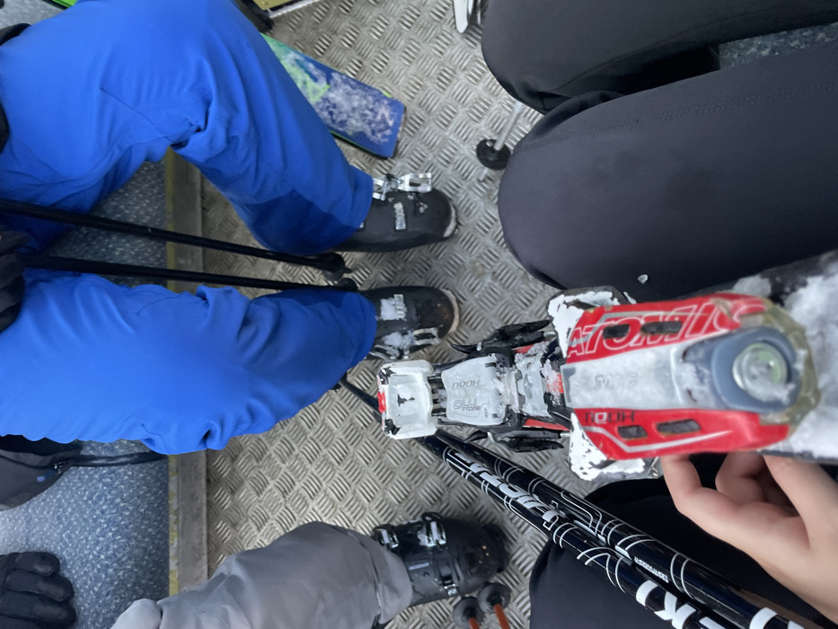 The image captures a high-angle view of multiple individuals and their ski equipment on a metal grated surface, likely within a ski lift or platform at a ski resort. The setting indicates cold, snowy conditions, with visible snow and ice clinging to the ground, ski boots, and bindings. Given the context, this scene is set in Courmayeur, Italy.
Several people are present, identifiable by their legs, boots, and a visible hand. On the left, a person wears vibrant blue ski pants and black ski boots, with black ski poles extending downwards. A portion of a ski with a blue and green pattern is visible behind their leg. In the mid-right, another person is wearing black ski pants. A hand, likely belonging to this person or another nearby, is seen holding the tip of a ski. At the bottom of the frame, a third person wears grey ski pants and black ski boots.
The ski equipment is a central focus. A prominent red and white ski binding, clearly branded "ATOMIC," is situated in the center-right. Adjacent to it is a white ski binding, partially obscured by snow and ice, with the text "DOOH" and "GULU PROME" visible. Black ski poles, potentially branded "LEKI" with white graphics, are crossed in the lower center. The metal grate flooring is typical of ski infrastructure, suggesting individuals are either waiting, taking a break, or in transit during a winter sports activity. The time of day appears to be daytime, with a somewhat subdued light, possibly overcast.