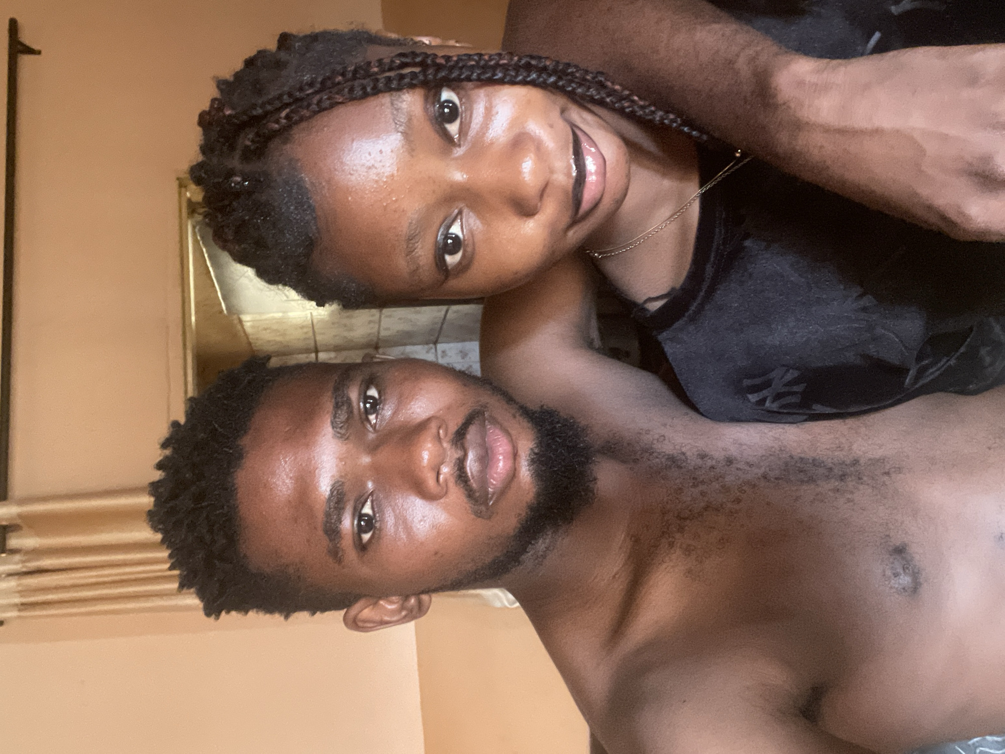 Two dark-skinned individuals pose for a selfie indoors. The woman, positioned towards the top of the frame, has braided hair and is wearing a dark top with a gold necklace. She is smiling. The man, located towards the bottom, has short, dark hair and a beard. He is shirtless. The background features a peach-colored wall, a partially visible mirror, and a curtain. The photo appears to be taken in a residential setting. There is no definitive evidence to confirm it was taken in Unguwar Gabas, Nigeria. The information about the location is not visually verifiable.