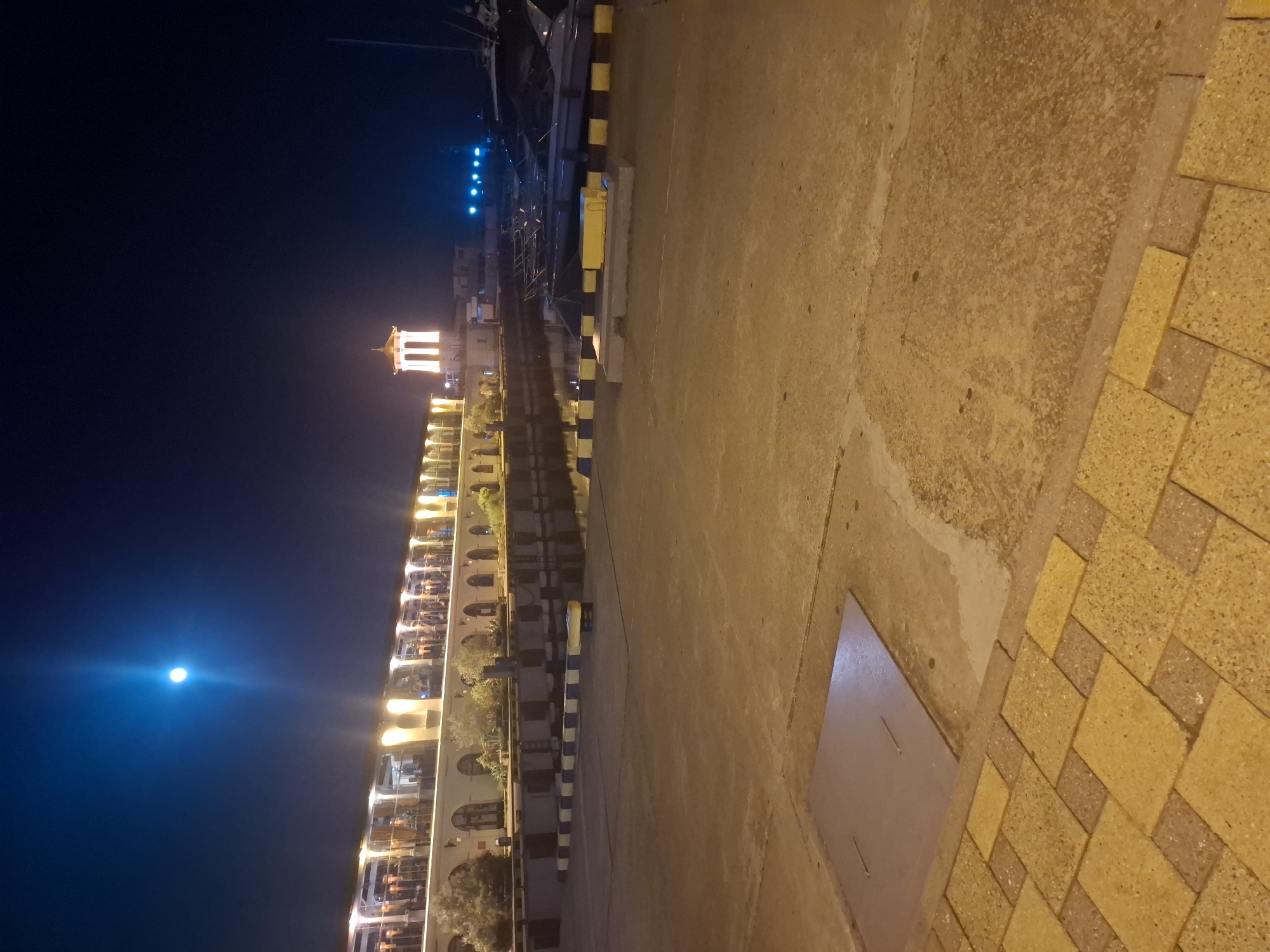 This image depicts a night scene at the Sochi, Russia, port or waterfront. A brightly lit building with arches and balconies lines the water's edge, suggesting it may house restaurants or shops. A distinctive white, illuminated tower stands out against the dark sky, which is dominated by a prominent full moon and a bright halo around it. In the foreground, there is a concrete ground, part of which is paved with yellow and brown rectangular tiles. The water reflects the lights from the building and the moon. Moored boats are visible in the middle ground, and a few blue lights dot the distant water. The scene conveys a sense of calm and illumination in a waterfront urban environment at night.