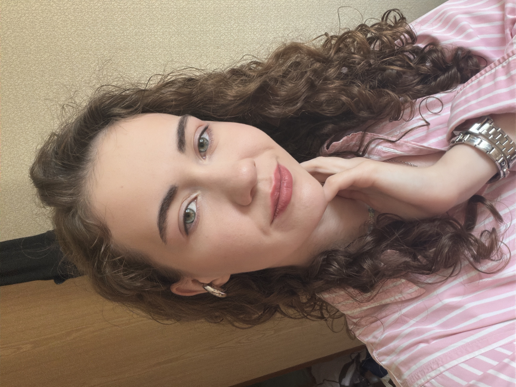 The image is a selfie of a young woman with curly brown hair and green eyes. She is wearing a pink and white striped shirt, silver earrings, and a silver watch. Her right hand is resting on her chin. The background appears to be a room with a wooden surface and textured wallpaper. There is no indication of the location being in Zavodoukovsk, Russia.