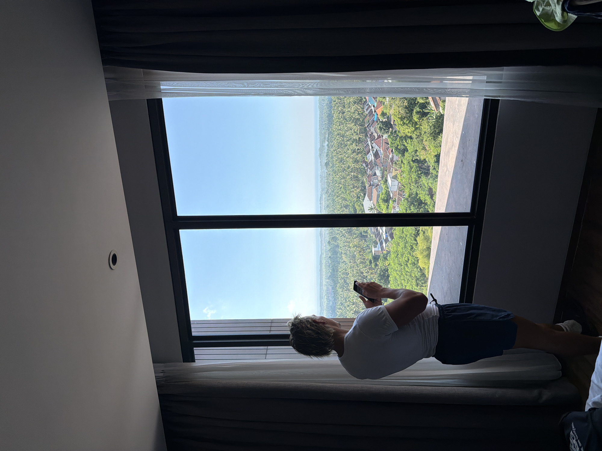 A blond-haired person wearing a white t-shirt and dark shorts stands in front of a large window, looking out at a landscape. The person holds a black smartphone in their right hand. Through the window, a dense jungle of palm trees stretches out, with a small village nestled among them. The village has buildings with red-tiled roofs and some white structures. In the distance, the ocean is visible under a clear blue sky. The window is framed by dark curtains on the sides and sheer white curtains on the right. The floor appears to be made of wood. The scene is lit by natural daylight coming through the window.
