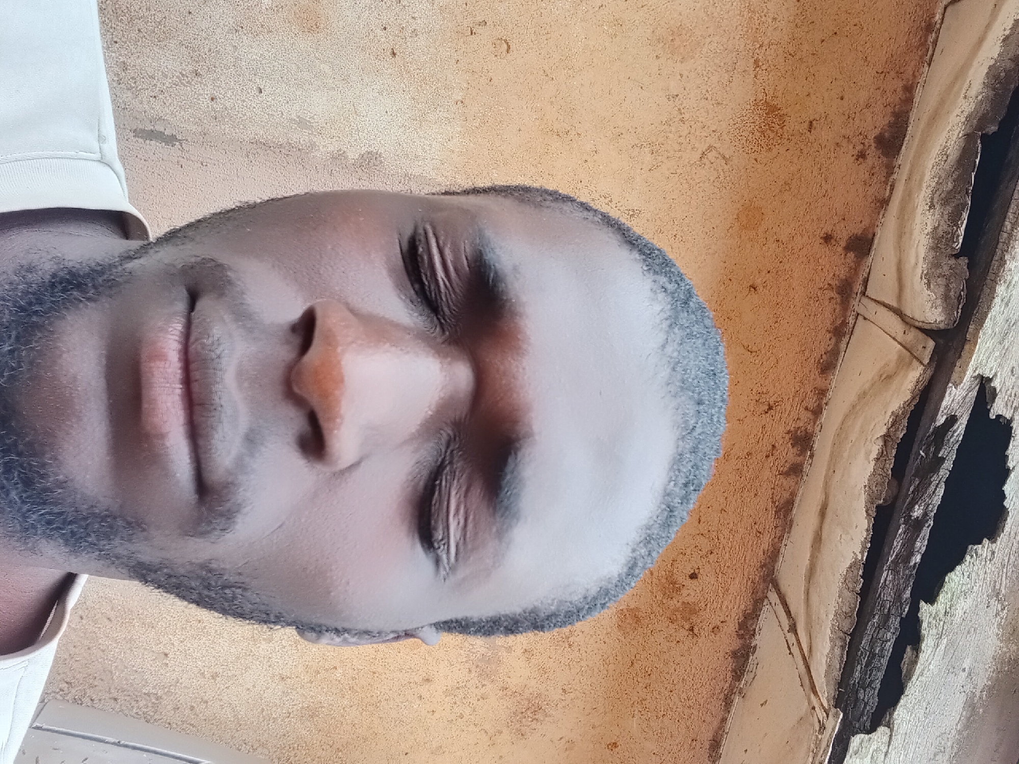 The image is a close-up, downward-angled selfie of a Black man with short hair and a beard. His eyes are closed, and he is wearing a light-colored t-shirt. The background consists of a textured, tan-colored wall with some discoloration and a damaged wooden structure overhead. The lighting appears to be natural, casting soft shadows on the man's face. The scene is outdoors, suggesting an urban environment. The location is identified as Bukuru, Nigeria.