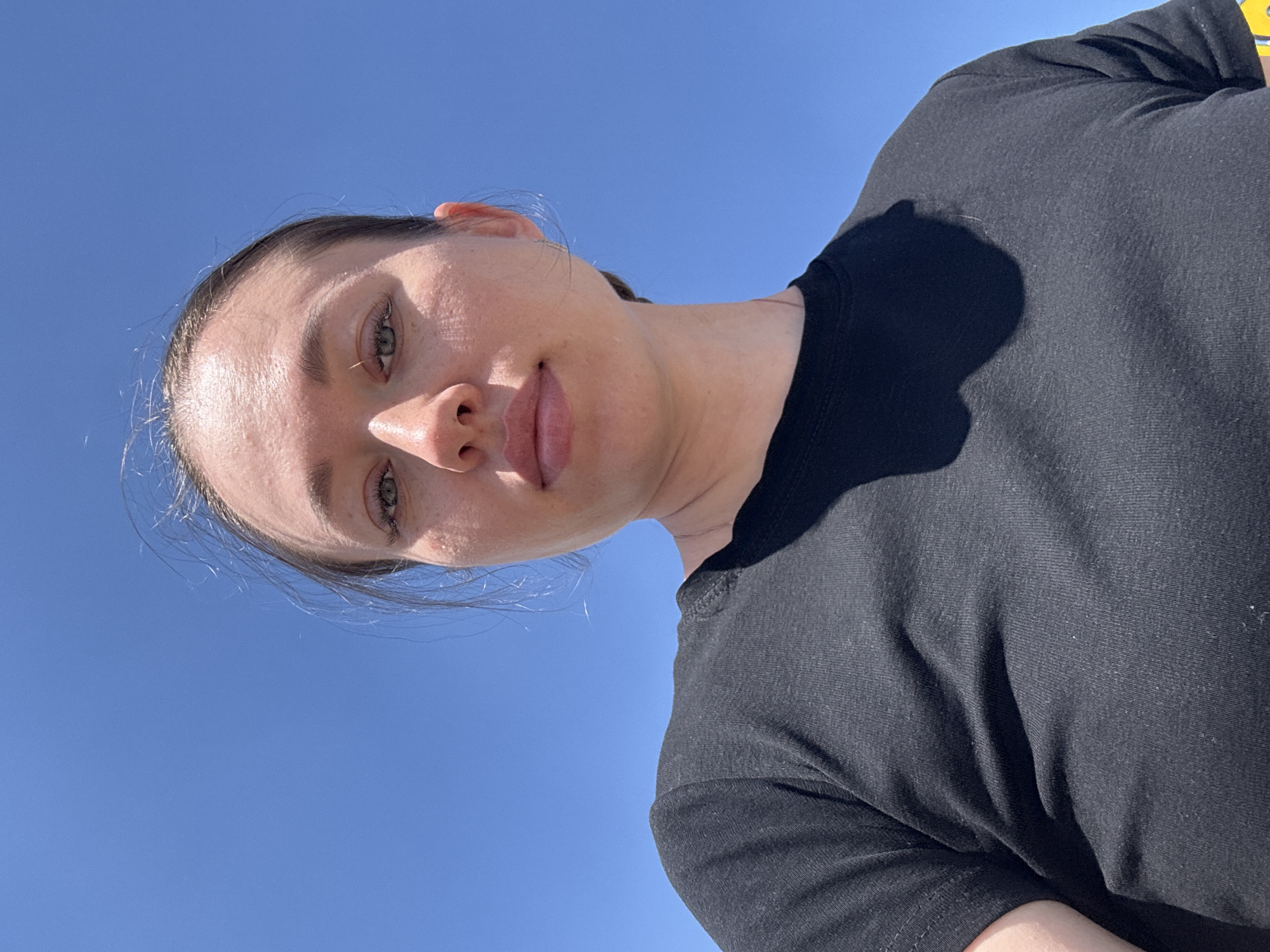 A woman is positioned in the lower left of the frame. Her face is tilted towards the viewer. She has light skin, brown hair pulled back, and green eyes. She's wearing a black t-shirt. The background is a clear blue sky. The location cannot be determined from the image.