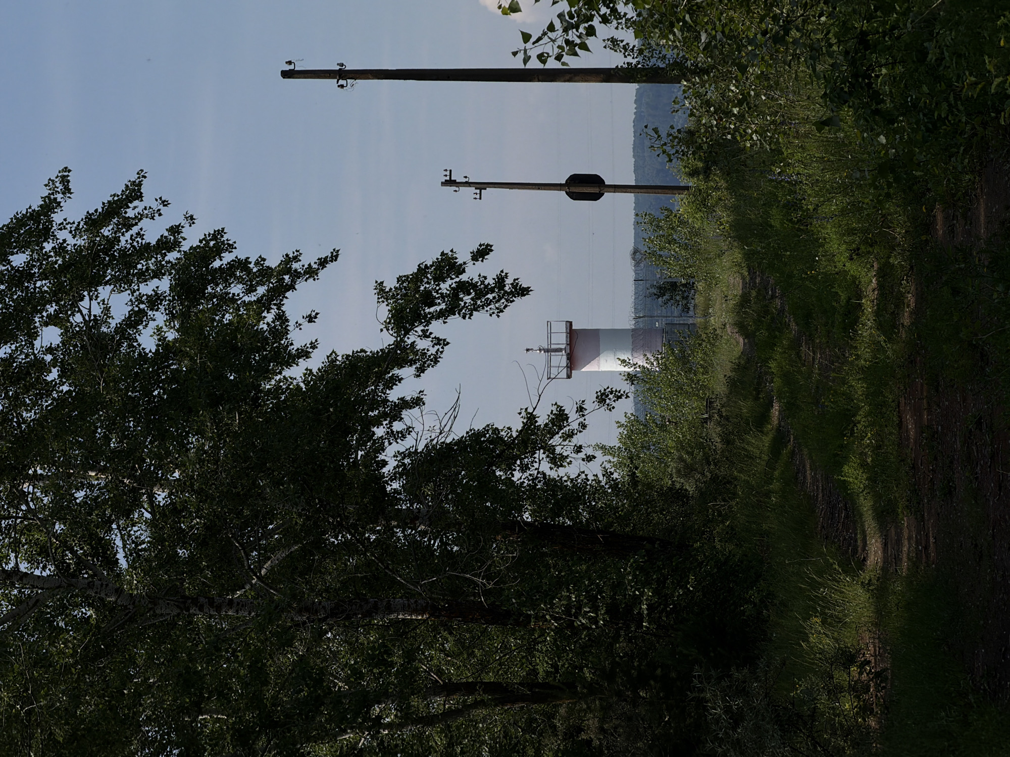 This is an outdoor, eye-level, medium shot photograph taken on a sunny day. The scene depicts a dirt path leading through lush green vegetation and trees. In the background, a red and white cylindrical structure, possibly a lighthouse or tower, is visible. To the right of the path, two tall utility poles stand, with one of them supporting a dark, oval-shaped object. The sky is clear and blue, with a few faint clouds. The overall impression is one of a natural, somewhat remote location within or on the outskirts of the city of Chaikovsky, Russia.