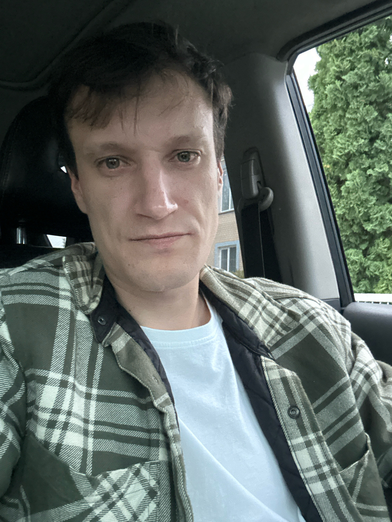 The image shows a close-up of a man sitting in a car, with his upper body and face visible. He is wearing a light blue t-shirt underneath a green and white plaid jacket. The man has short, dark hair and light-colored eyes. He is looking directly at the camera with a neutral expression. The interior of the car is visible, including part of the headrest and a seatbelt. Through the car window, a dense green tree is visible, along with a building with windows. The image is taken from a slightly elevated angle.