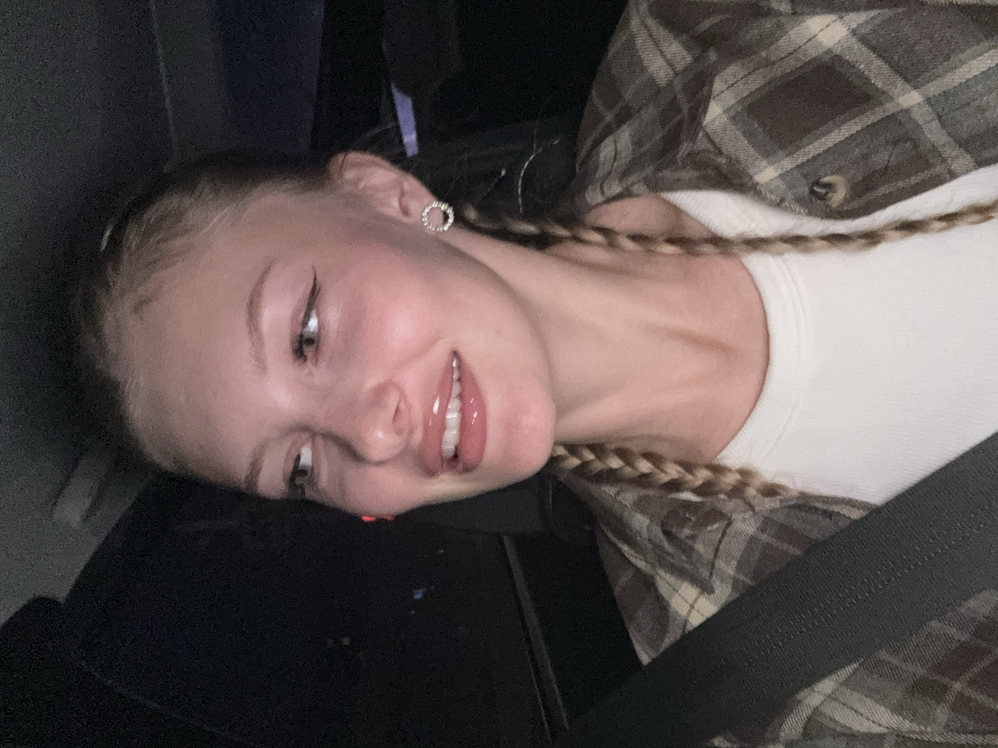 A young woman with blonde hair styled in two braids is seated in the passenger seat of a vehicle at night. She is wearing a brown and white plaid shirt over a white tank top and a black seatbelt is visible across her chest. She is looking to her left with her mouth slightly open, revealing her teeth. Her makeup includes winged eyeliner and lip gloss. She is wearing a circular, jeweled earring in her right ear. The background is dark, suggesting an urban setting at night, possibly on a road within the city of Perm, Russia.