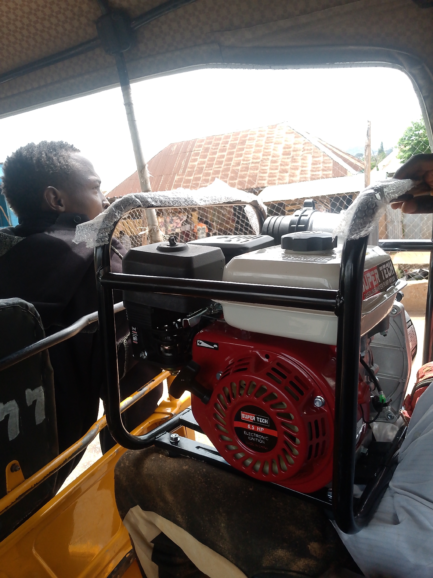 The photograph shows the interior of a yellow vehicle, possibly a taxi or auto-rickshaw, in Bukuru, Nigeria. A generator with a red engine is placed inside the vehicle. The generator, labeled "SUPER TECH" with "6.5 HP" and "ELECTRONIC IGNITION MADE IN CHINA", is secured within a black metal frame. A man sits beside it, looking out the window. Outside, there are glimpses of buildings with corrugated iron roofs. The handle of the generator's frame is wrapped in bubble wrap.
