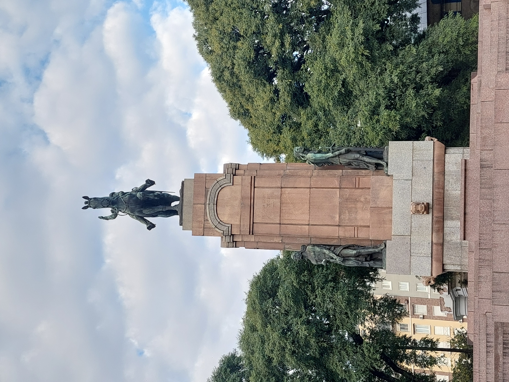 The image displays a prominent equestrian monument, likely situated in a public space in Buenos Aires, Argentina. At the apex of a tall, multi-tiered pedestal stands a dark metal statue of a military figure on horseback, with the horse depicted in a dynamic, possibly rearing, pose. The rider is in military attire.
The substantial pedestal is constructed from reddish-brown and greyish stone, featuring classical architectural elements such as cornices and decorative carvings. Several smaller, allegorical bronze figures are positioned around the base of the pedestal, adding to its commemorative nature.
Lush, dense green trees frame the monument on both sides and form a verdant backdrop. Partially visible behind the foliage and the monument are light-colored urban buildings with multiple windows, indicating an urban setting. The sky above is partly cloudy, suggesting it is daytime, likely late morning or early afternoon.
Visible inscriptions on the stone pedestal provide historical context, including the dates "1810", "1813", "BATALLA DE SUIPACHA", "1816", and "1817". These details confirm the monument's historical significance, specifically referencing the Battle of Suipacha, a key event in the Argentine War of Independence. The monument likely honors a hero or event from this period.