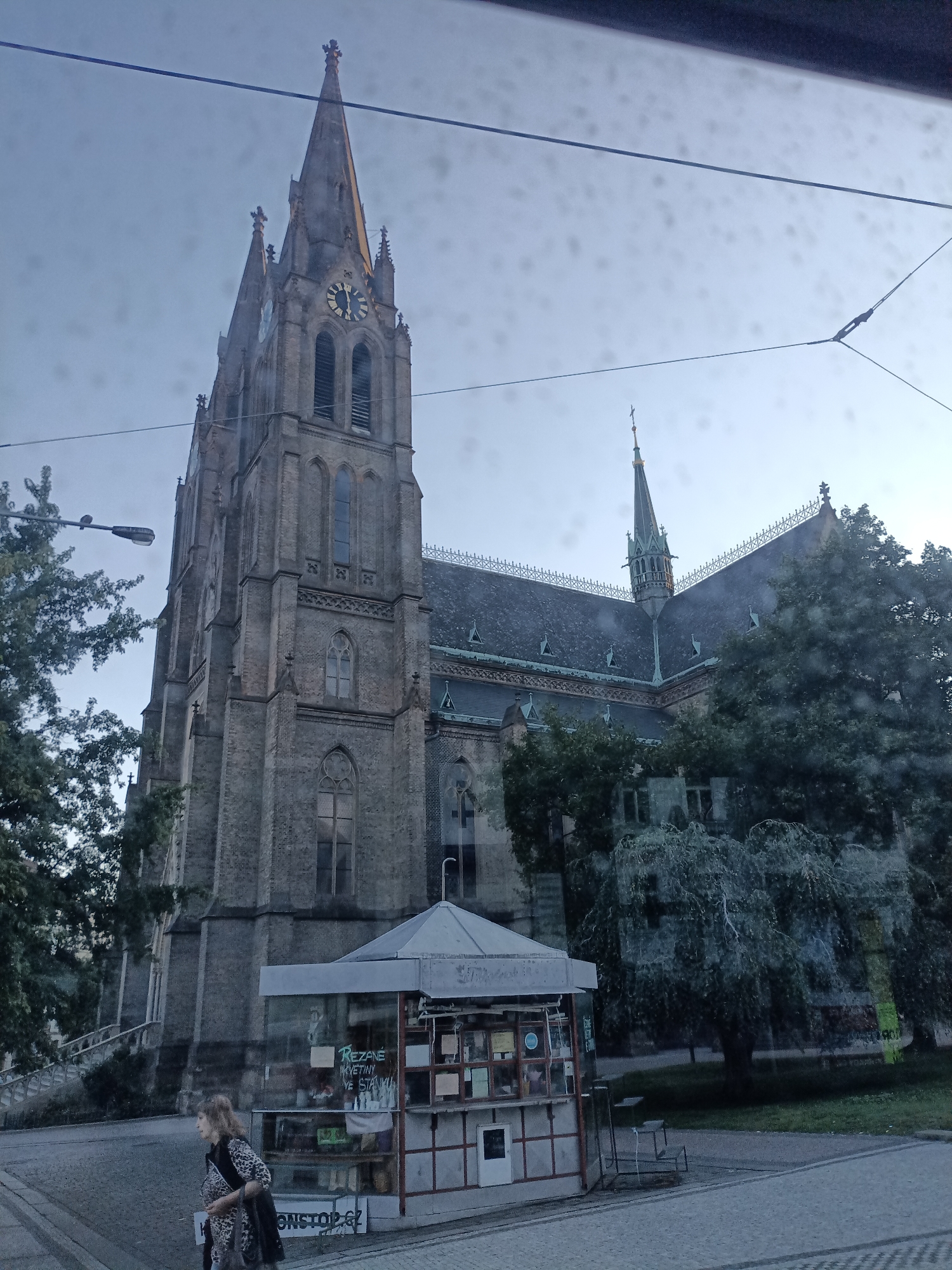 The image shows the exterior of the Church of Saints Peter and Paul in Vyšehrad, Prague, Czechia. The church, constructed primarily of brick, features a prominent clock tower with a spire and ornate detailing. In the foreground, a small kiosk is visible, displaying various items for sale through its glass windows. A woman with blonde hair, wearing a leopard-print top and a dark jacket, walks past the kiosk. The scene is set on a cobblestone street with trees and overhead power lines present in the environment. The sky is overcast, suggesting it might be late afternoon or early evening.