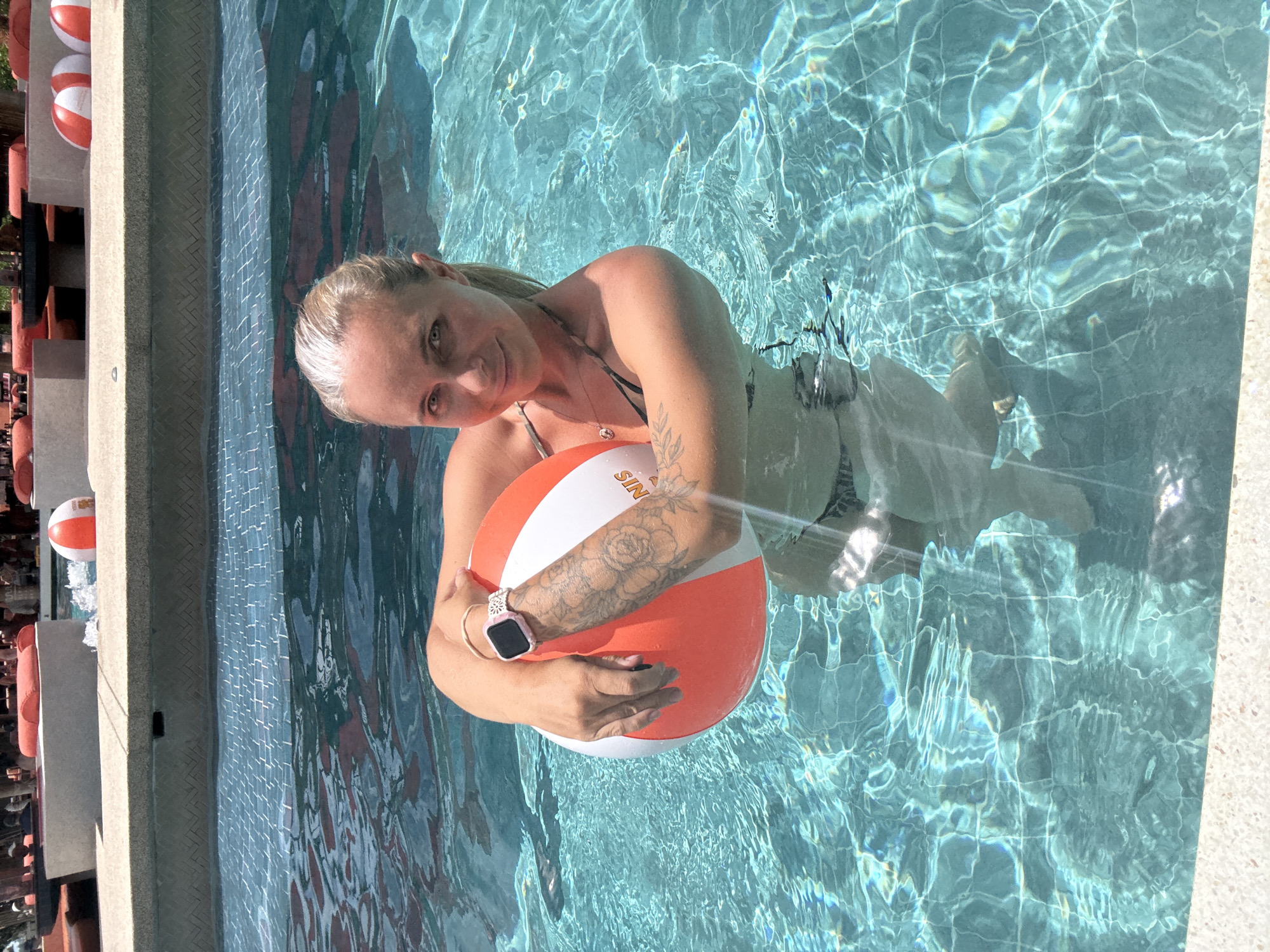 A woman with blonde hair pulled back in a ponytail, wearing a dark patterned bikini, is in a swimming pool holding a striped orange and white beach ball. She has a tattoo of roses on her left forearm and is wearing a pink smartwatch. The pool has turquoise water with sunlight reflecting off the surface, creating a grid pattern from the tiles below. In the background, out of focus, are lounge chairs with orange cushions and some people. The location is identified as Ko Samui District, Thailand.