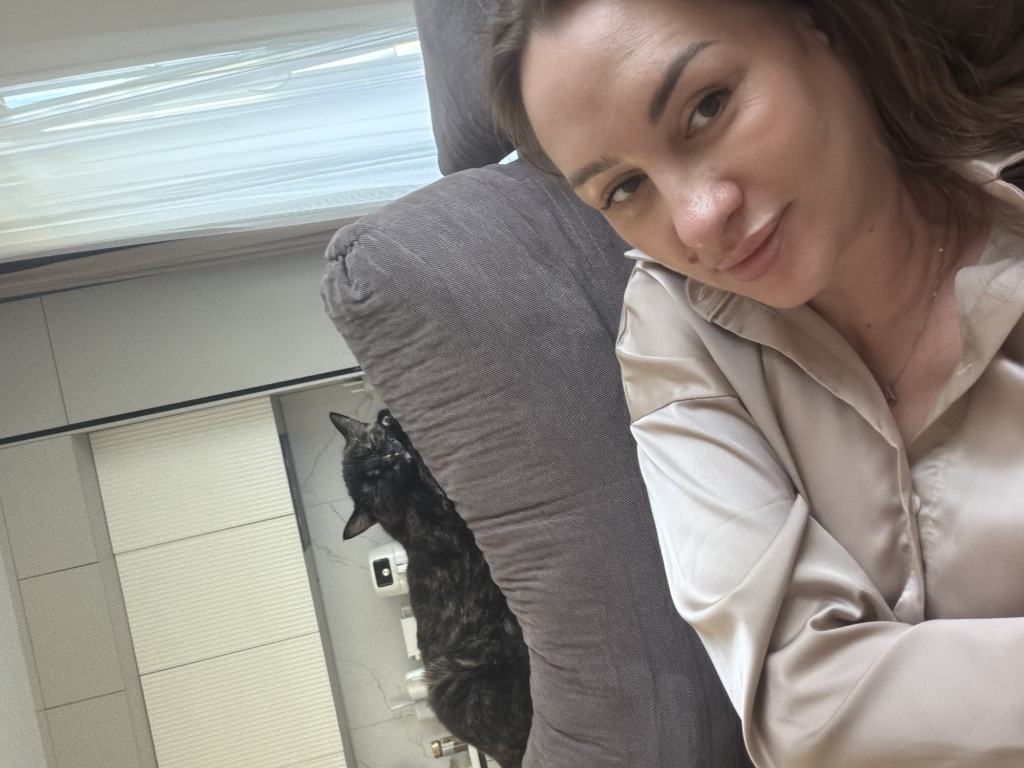 A woman is taking a selfie in a room. She is wearing a light-colored satin shirt and has brown hair. She is looking towards the camera and smiling slightly. Next to her, partially obscured by a gray couch, is a tortoiseshell cat. The cat is looking upwards with its mouth slightly open. In the background, there is a window with white blinds and a portion of a wall that appears to be tiled. The lighting suggests it could be daytime. The overall scene appears to be indoors, possibly a living room or bedroom. There is no visible text in the image.