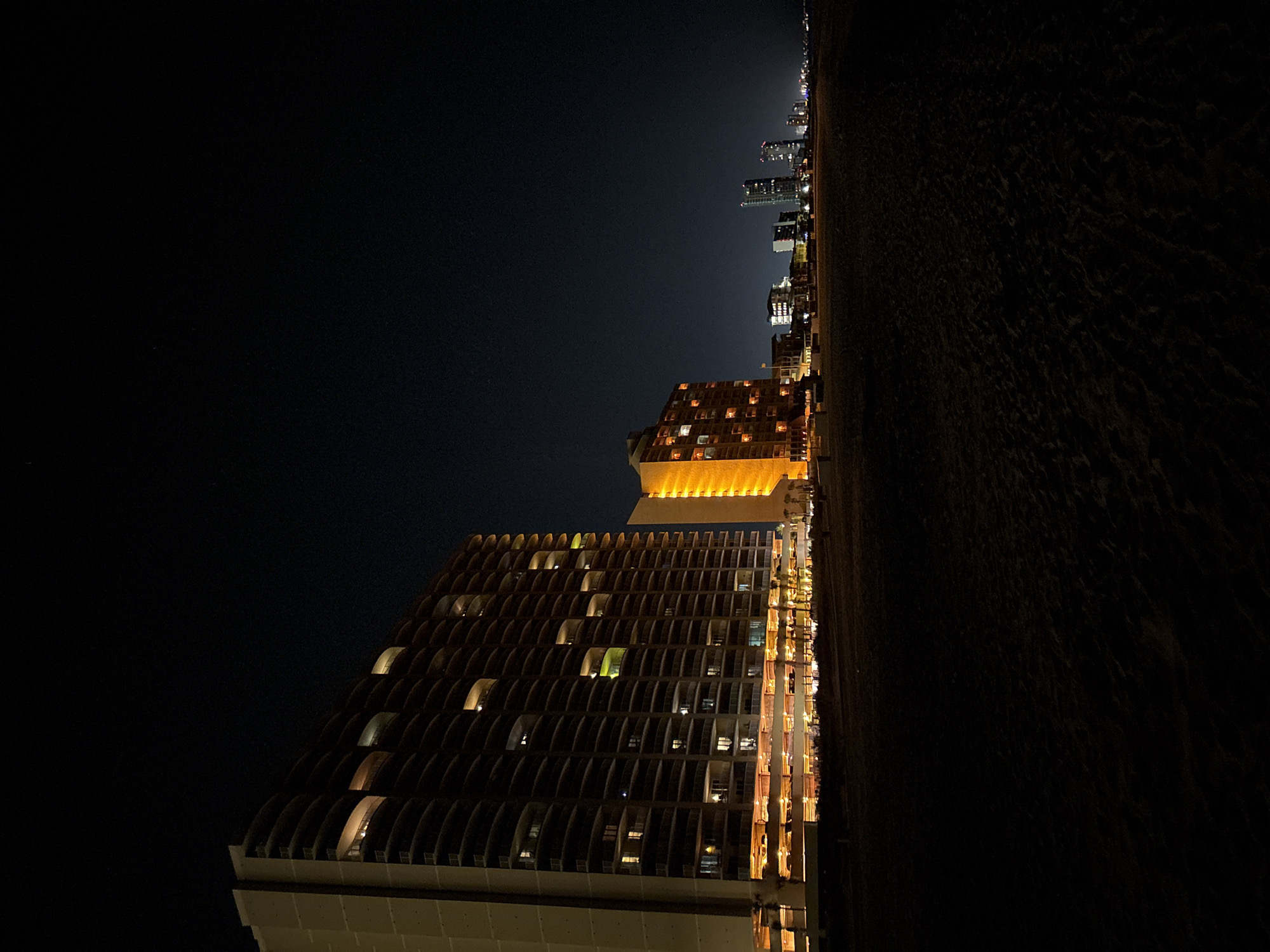 The image depicts a nighttime scene in Panama City Beach, Florida, United States of America. It shows a dark beach and the skyline of a city, including several buildings that are lit up. The foreground features two illuminated high-rise buildings. The top of the image shows a dark sky. The beach is dark. The angle is vertical and oriented in a way that rotates the viewer's head to view it.