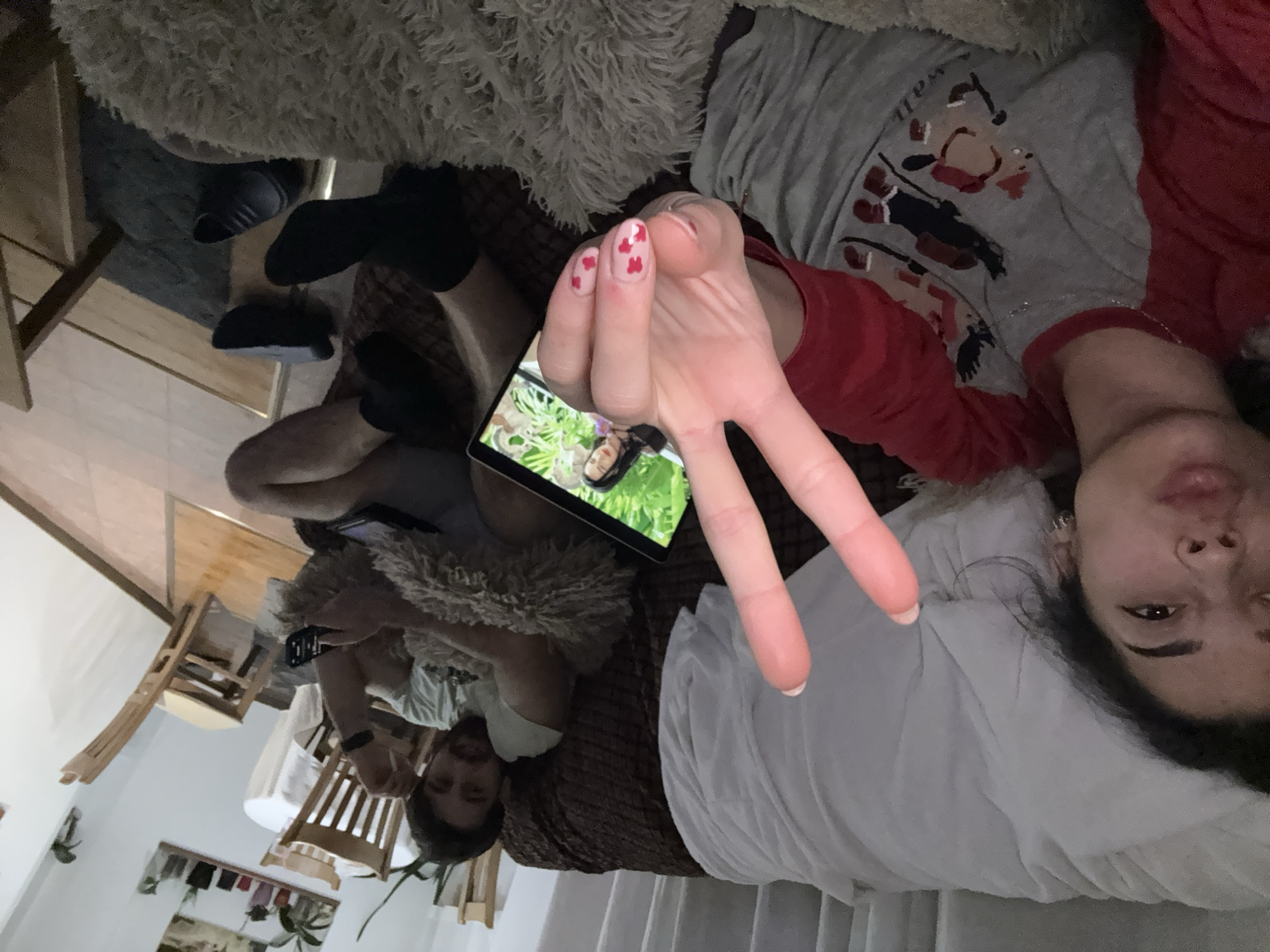 The image depicts an interior setting, possibly in Sofia, Bulgaria. Two people are visible, seemingly in a relaxed or reclining position. One person is in the foreground, near a pillow, with a hand making a "peace" sign. The nails of this hand are decorated with small red hearts. The second person, located further back, appears to be watching something on a tablet. Furniture such as a sofa, chairs, and a small table are present. A furry blanket or throw is draped over some of the furniture. There is a glimpse of clothing hanging on a rack in the background, and possibly a plant. The lighting is subdued.