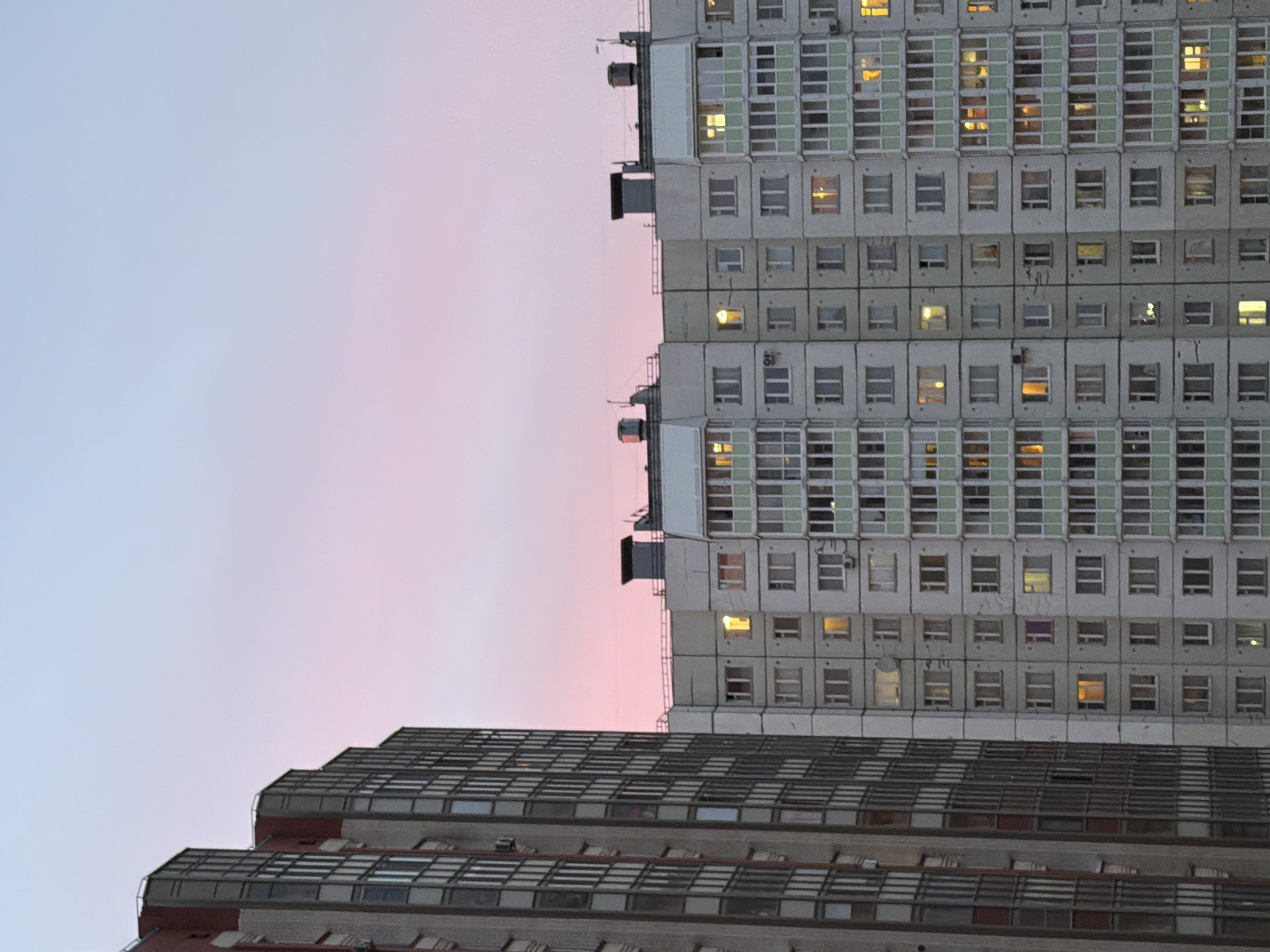 The image depicts a low-angle view of apartment buildings in what appears to be Saint Petersburg, Russia, during twilight. The sky transitions from a pale blue to a pink hue. The taller building on the right is a grey high-rise apartment block with a grid of windows, many of which are illuminated. To the lower left is a tiered building with a similar grid-like window pattern and a red brick base. The overall composition suggests a typical urban landscape at dusk in Saint Petersburg.