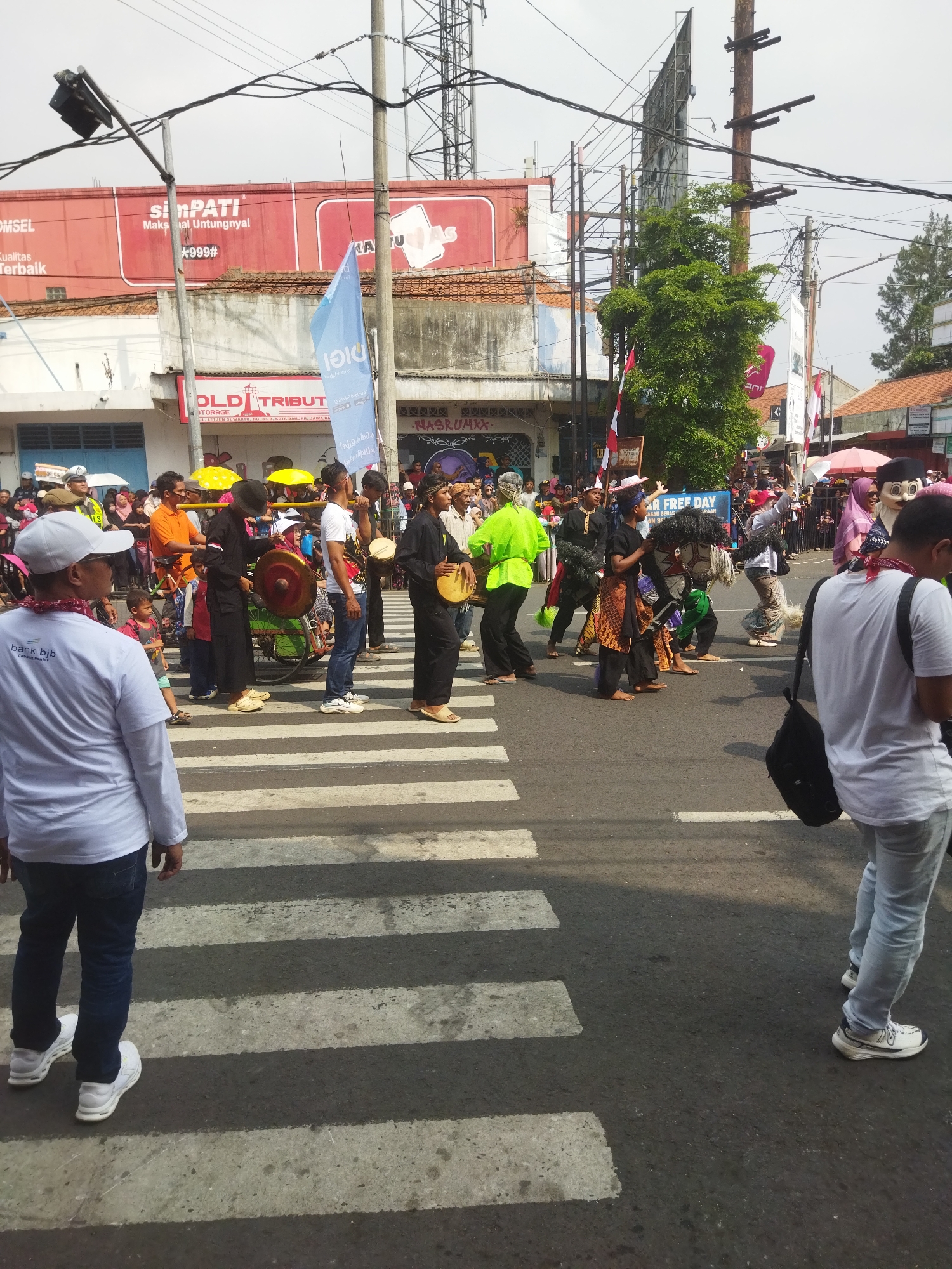 In the city of Banjar, Indonesia, a festive procession is underway on a street, marked by a pedestrian crosswalk. A group of individuals in traditional attire are participating, some playing musical instruments. One person in a black outfit is playing a large gong attached to a green wheeled cart, while another in a similar outfit is playing a smaller drum. In the foreground, two men stand on the crosswalk. The man on the left wears a white long-sleeved shirt with a red bandana around his neck and a white cap, while the man on the right wears a white t-shirt, blue jeans, and a black bag slung over his shoulder. Behind them, a crowd of onlookers lines the street, many holding umbrellas and flags. Buildings with red and white facades are visible in the background, along with utility poles and power lines. The sky is overcast, suggesting a daytime event.