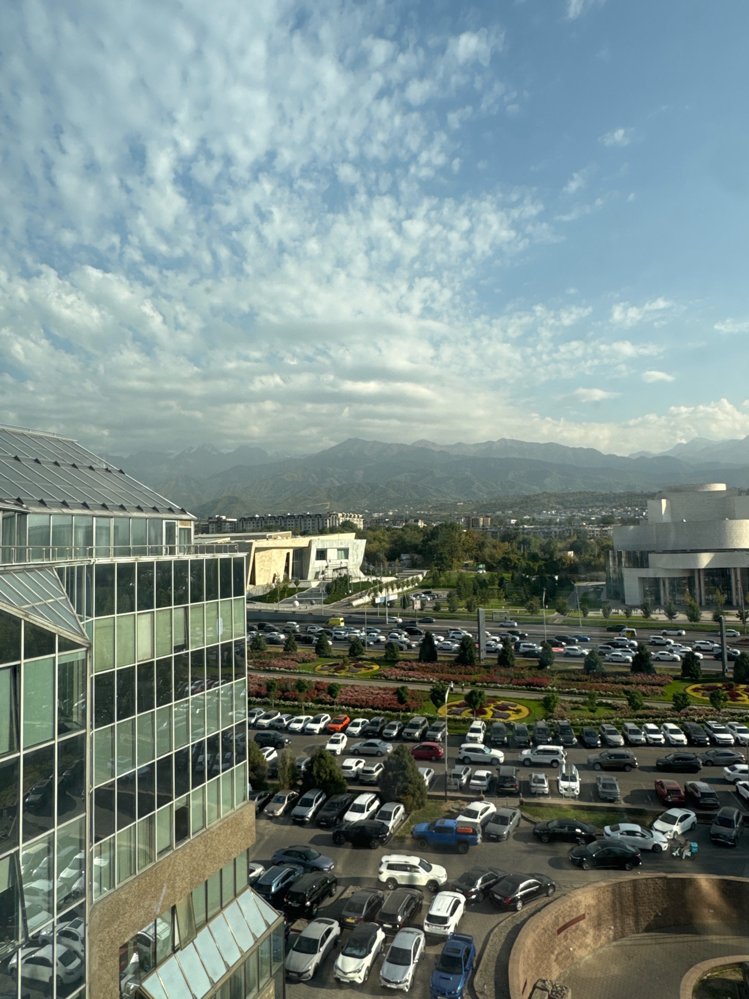 This is an aerial view of a modern city district in Almaty, Kazakhstan, with a focus on a large, multi-level parking lot filled with numerous cars and several modern buildings. The foreground features a glass-sided building with reflective windows, showing partial reflections of cars and the sky. Beyond this building, a vast asphalt parking area stretches across the frame, occupied by a dense collection of various vehicles, including sedans, SUVs, and vans, in a range of colors.
Adjacent to the parking lot are several contemporary architectural structures, including a building with a curved facade and a flat roof, and a lower, rectangular building with a light-colored exterior. Green spaces with manicured flower beds and trees are interspersed between the buildings and the parking areas, adding a touch of nature to the urban landscape.
In the background, the cityscape extends towards a range of majestic, hazy mountains under a bright blue sky dotted with scattered white clouds. The lighting suggests it is daytime, possibly late morning or early afternoon, with sunlight casting shadows and highlighting the different textures and surfaces. There are no people visible in the scene. The overall impression is one of a bustling urban environment integrated with natural mountainous scenery.