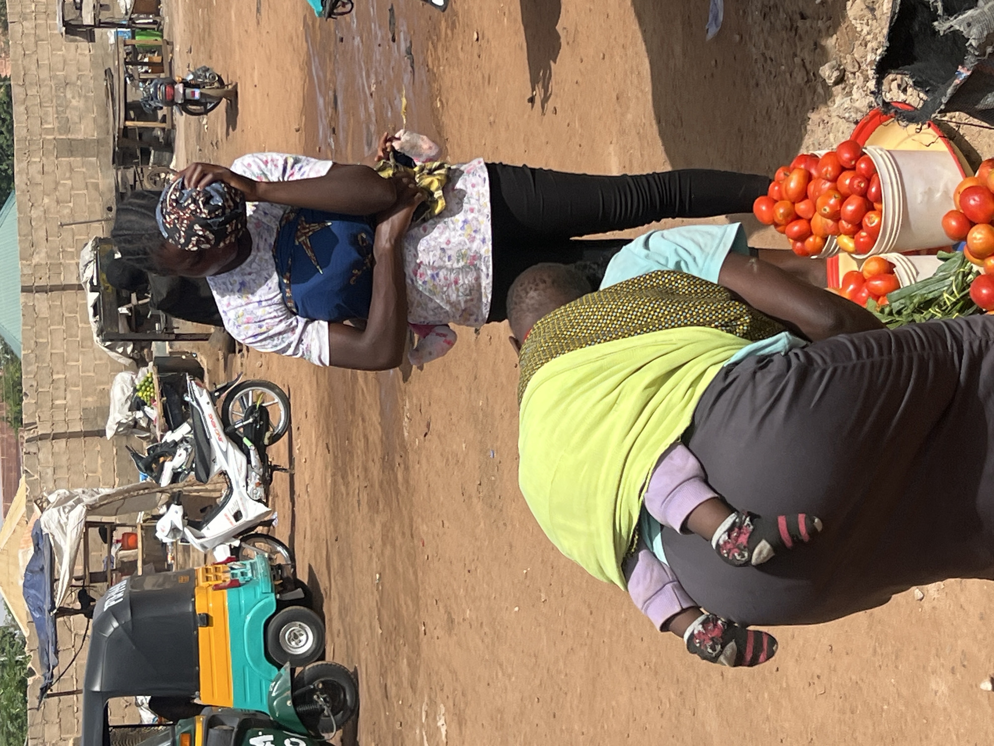 This media file captures an outdoor market scene under bright daylight, likely in Gura Top, Nigeria. The ground is unpaved and dusty.
In the foreground, two women are visible. To the left, a woman with dark skin wears a patterned headwrap, a light patterned shirt, and dark trousers. She is looking down, with her hands clasped. To the right, another woman, seen from behind, is carrying a baby on her back in a sling or wrap. She wears a bright lime-green garment over a darker top and dark trousers. The baby's legs, wearing light purple pants and dark, patterned socks, are visible, extending from the wrap.
Behind these individuals, there are several white plastic buckets overflowing with ripe red tomatoes, suggesting a market stall. Some green leafy vegetables are also present. In the background, there's a simple structure with a partial concrete block wall. To the far left, a yellow and blue/green auto-rickshaw (tuk-tuk) and a white motorcycle are parked, indicating a busy area.
The strong sunlight creates distinct shadows, characteristic of a sunny day. No clear text is visible on any objects or clothing. The overall atmosphere appears to be a regular, bustling market environment.