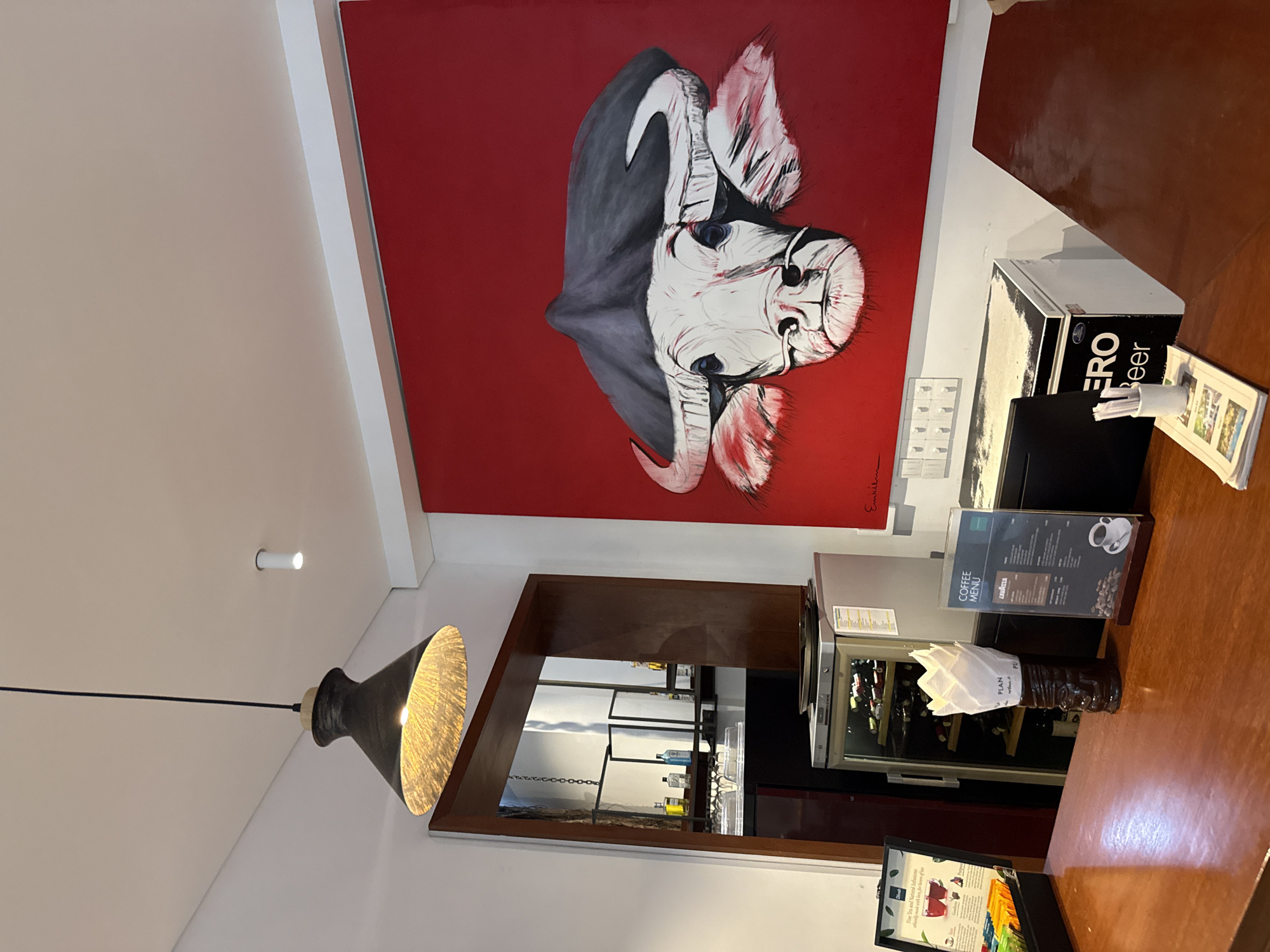 The image depicts the interior of a cafe or bar, potentially located in Khan Daun Penh, Cambodia. A prominent feature is a large, vibrant red canvas displaying a stylized painting of a bull's head. The artwork is mounted on a white wall.
In the foreground, a polished wooden counter is visible. On the counter, to the left, sits a black box containing various tea sachets, with text indicating "Fine Tea and Natural Infusions." To the right of the tea box is a dark brown, tiki-style holder filled with white napkins. Further to the right, a small, clear stand displays a "COFFEE MENU" with offerings including "LAVAZZA." Below the menu, a ceramic cup and saucer are illustrated.
Behind the counter, a compact silver beverage cooler is present, with its glass door revealing rows of wine bottles. Adjacent to the cooler is a white chest with a black lid, featuring large white text that reads "ZERO Beer." A computer monitor is also positioned on the counter.
Above the counter, a dark, cone-shaped pendant light with a textured interior hangs from the ceiling. A small white cylindrical light fixture is also visible on the ceiling in the background. To the left, a wooden framed opening reveals a glimpse of a bar area with shelves, glassware, and bottles. The overall lighting suggests an indoor ambiance, possibly with artificial light sources. No people are visible in the image.