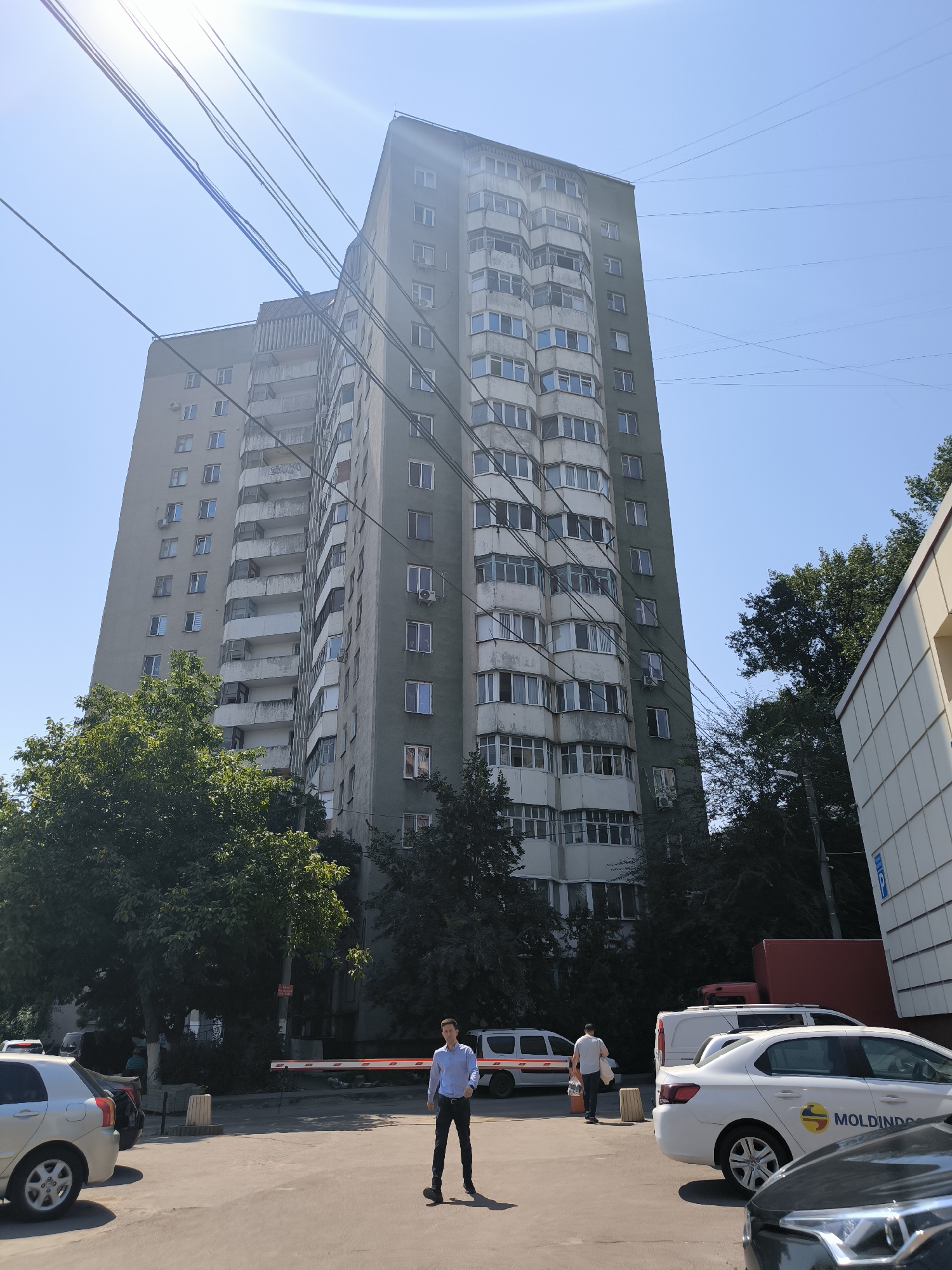 The image depicts a street scene in Chișinău, Moldova, dominated by a tall, grey apartment building with numerous balconies and windows. Overhead, a tangle of electrical wires stretches across the bright blue sky. In the foreground, a paved area serves as a parking lot with several cars. A white sedan with the "MOLDINDO" logo is parked on the right. On the left, a silver car is visible. A man in a light blue shirt and dark pants walks towards the right, and another person, seen from the back, walks towards the left carrying bags. A red and white barrier arm is lowered across the street. Green trees are interspersed with the buildings. The sun casts strong shadows, indicating a clear, sunny day.