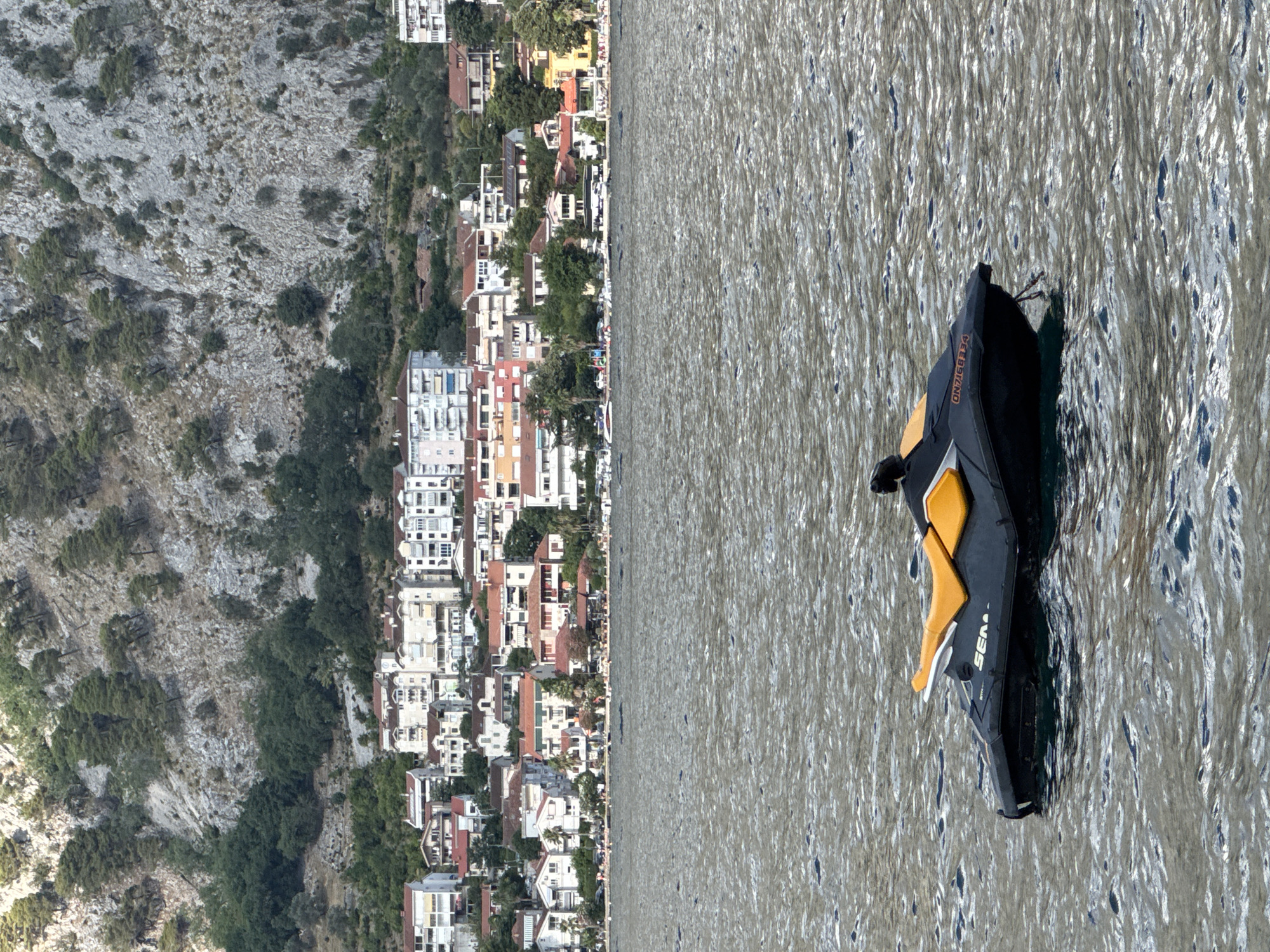 A black and orange jet ski floats on the rippling water of the Bay of Kotor, with the town of Prčanj, Montenegro, and a steep, rocky mountainside visible in the background. The jet ski, positioned in the lower center of the frame, has its orange seat slightly forward of the black body. The town of Prčanj, a coastal settlement, is characterized by a cluster of buildings with red and white roofs, nestled between the water and the imposing, tree-covered mountain. The mountain itself displays a rough, grey, rocky texture with sparse vegetation. The water exhibits a shimmering, sunlit surface with visible wave patterns. No people are discernible on the jet ski or in the immediate vicinity. The scene is captured from a water-level perspective.
