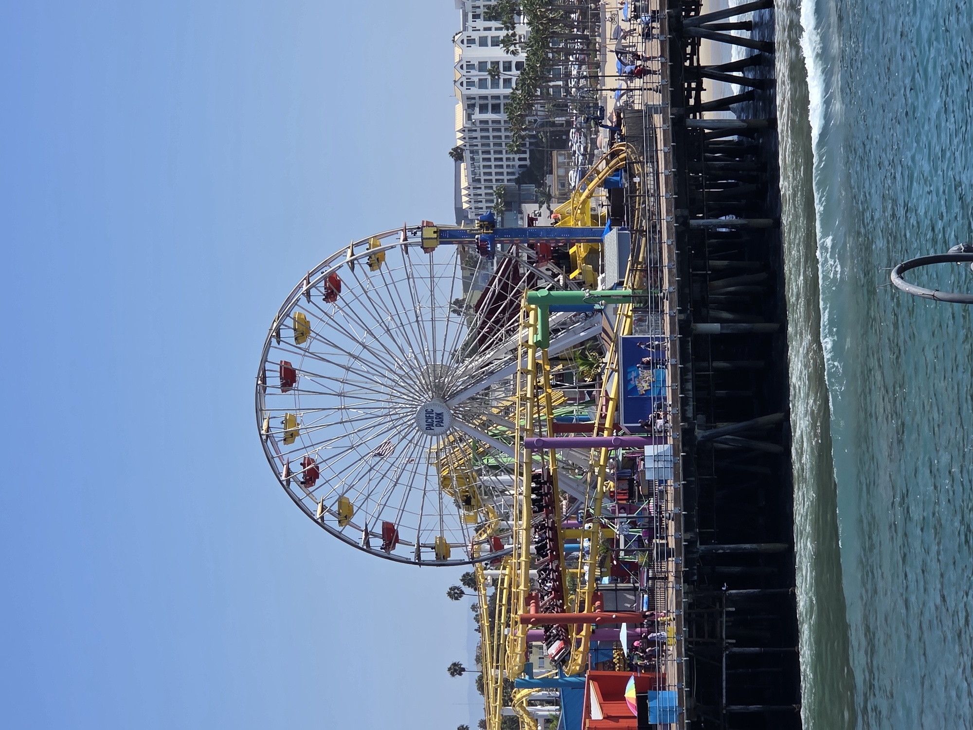 The image shows Pacific Park on the Santa Monica Pier in Santa Monica, California, USA. A large Ferris wheel with red and yellow gondolas is visible, along with a yellow roller coaster. The pier is crowded with people enjoying the amusement park. The ocean is in the foreground, with waves crashing against the shore. In the background, there are palm trees and buildings. The sky is clear and blue.