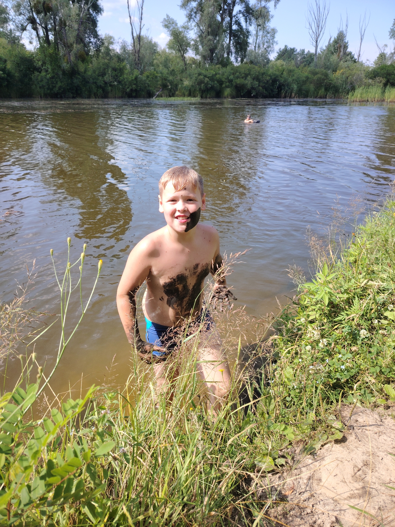 A young boy stands in a murky body of water, his body covered in dark mud. He is smiling widely, with mud smeared across his face, particularly around his eyes and mouth. The boy is wearing blue swimming trunks. The water is calm and reflects the surrounding trees and sky. The scene is framed by tall green grass and reeds along the bank on the right side of the image, with a sandy area visible at the very bottom. In the background, a dense line of trees forms the opposite bank of the water. The sky is bright blue, suggesting a sunny day. In the distance, another person is visible swimming in the water. The overall impression is one of playful, outdoor recreation on a warm day. The location context of Oseshchyna, Ukraine, suggests this is a natural setting, likely a river or lake. The time of day appears to be daytime, given the bright sunlight. There is no visible text in the image.