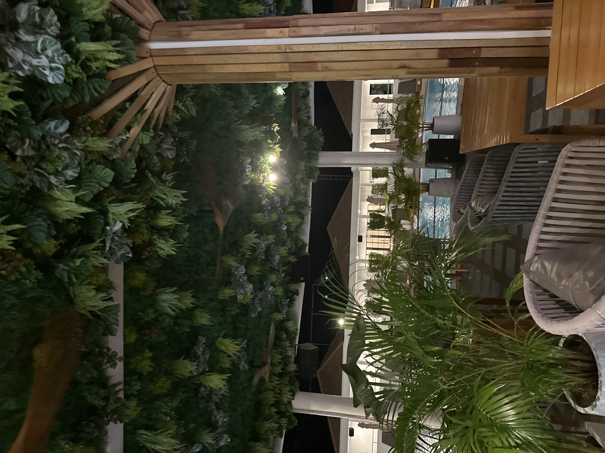 This is a photograph of an outdoor seating area at night, with a swimming pool in the background. The area is covered by an overhead structure that is heavily decorated with artificial foliage, creating a jungle-like atmosphere. In the foreground, there are several potted plants, including a large palm plant, and outdoor furniture such as chairs and tables made of wood and woven materials. The swimming pool is illuminated, and lounge chairs are visible around its perimeter. In the background, there are buildings with red-tiled roofs. The lighting suggests it is evening or nighttime. There are no people visible in the image.