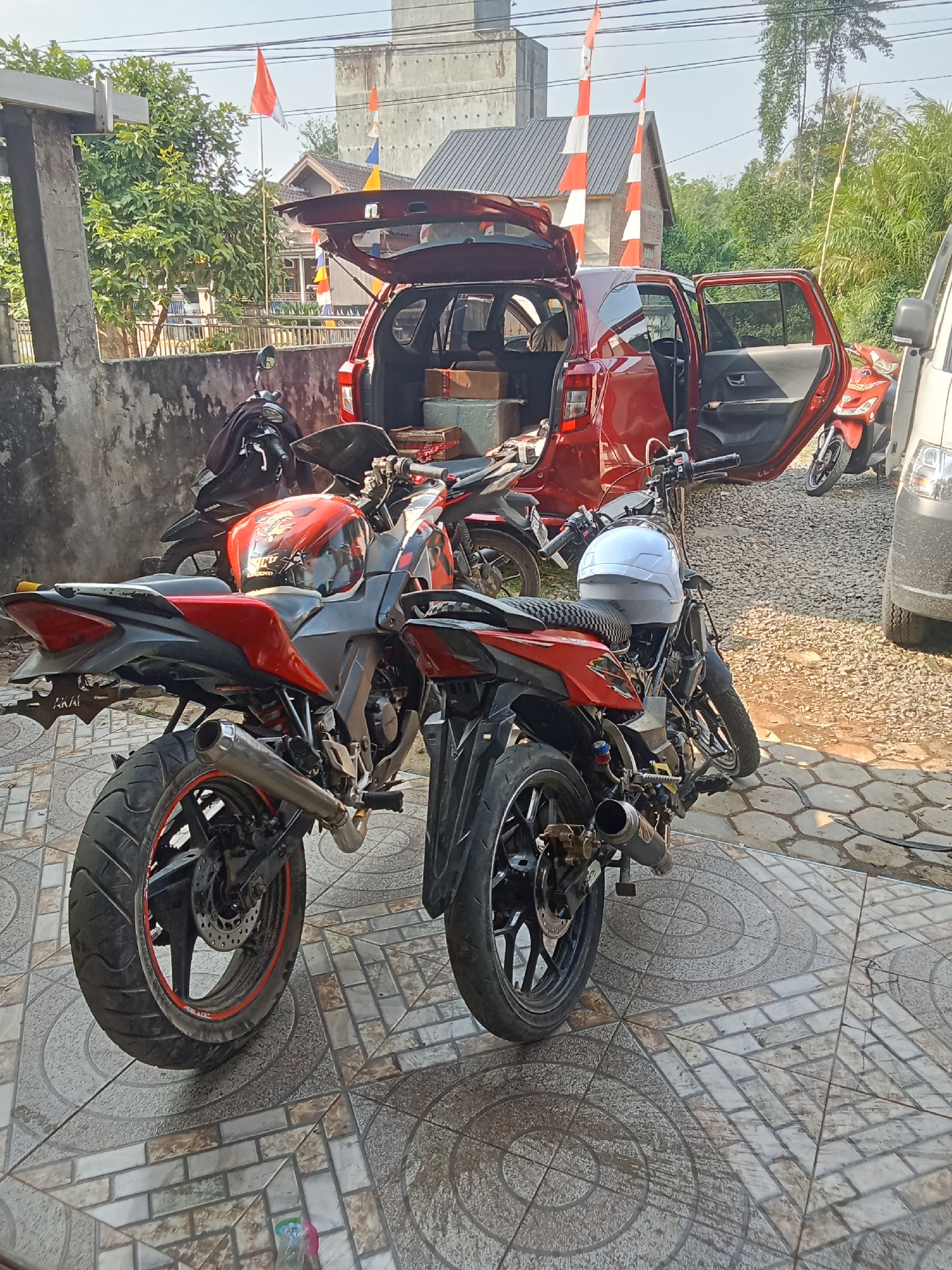 In Jirak, Indonesia, two motorcycles, a red and black sports bike and a black underbone motorcycle, are parked on a tiled patio in front of a red car with its trunk and driver's side door open. The car appears to be loaded with boxes. The scene is outdoors, with a wall to the left and lush greenery and buildings in the background. Several red and white striped flags are visible, indicating a possible celebration or event. A white motorcycle helmet rests on the seat of the black underbone motorcycle. The overall impression is one of a busy, potentially transitional moment, with items being loaded or unloaded from the vehicle.