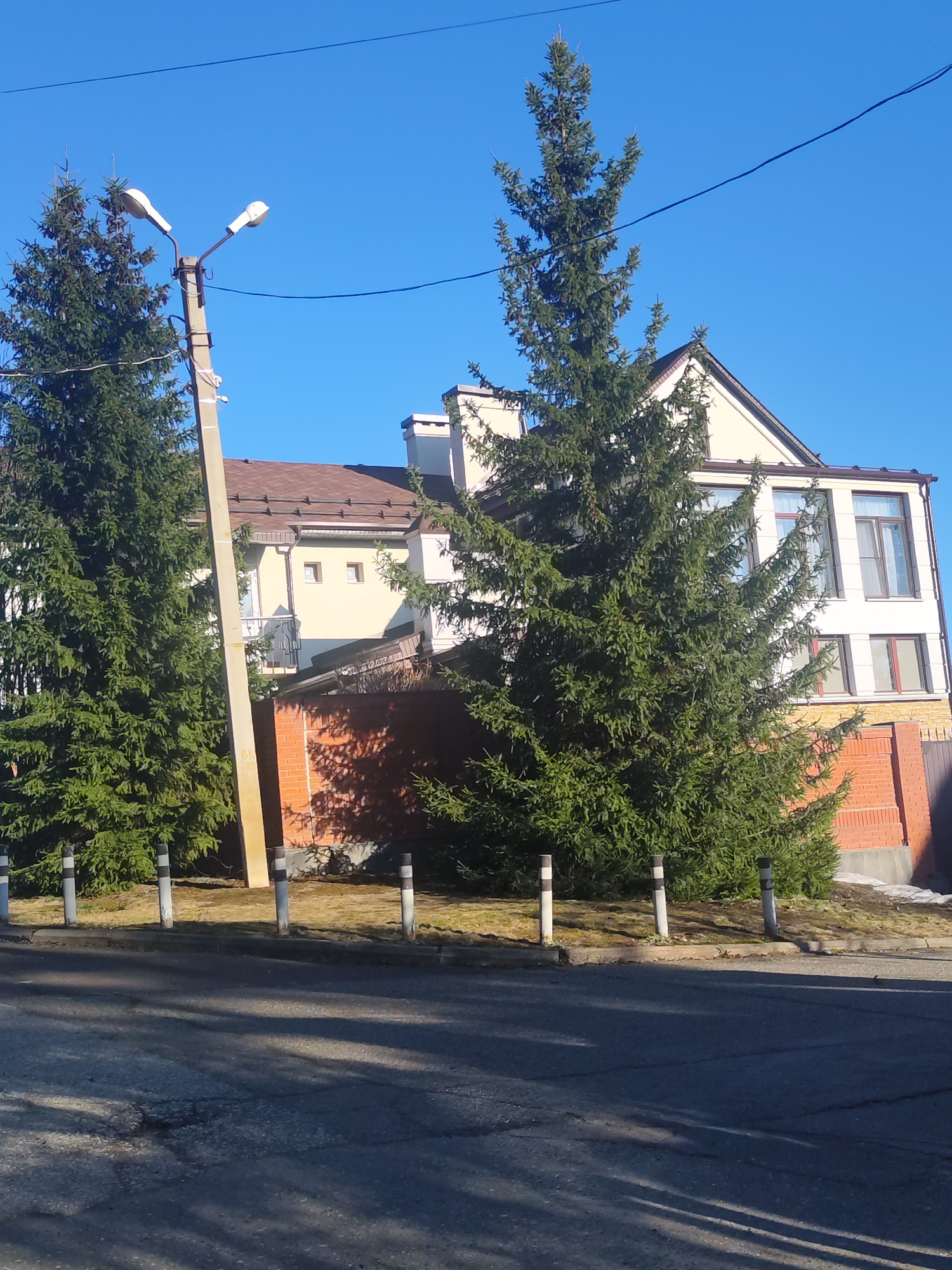 The image depicts a street scene in what is presumed to be the city of Прикамский, Russia. On the left side of the frame stands a tall, slender conifer tree positioned next to a leaning light post with two lamps. Adjacent to the tree, there is a series of short, cylindrical posts alternating black and white. Behind the posts, a two-story building is partially visible. The building features light-colored walls and a brown roof, with architectural details including a balcony, windows, and a chimney. A low brick wall extends from the building. The foreground is dominated by a street exhibiting cracked and uneven surfaces, suggesting it may be in need of repair. The sky is a clear, bright blue, indicative of a sunny day. Power lines stretch across the sky above the building and trees. There are no people visible in the frame. The overall impression is one of a quiet, suburban environment.