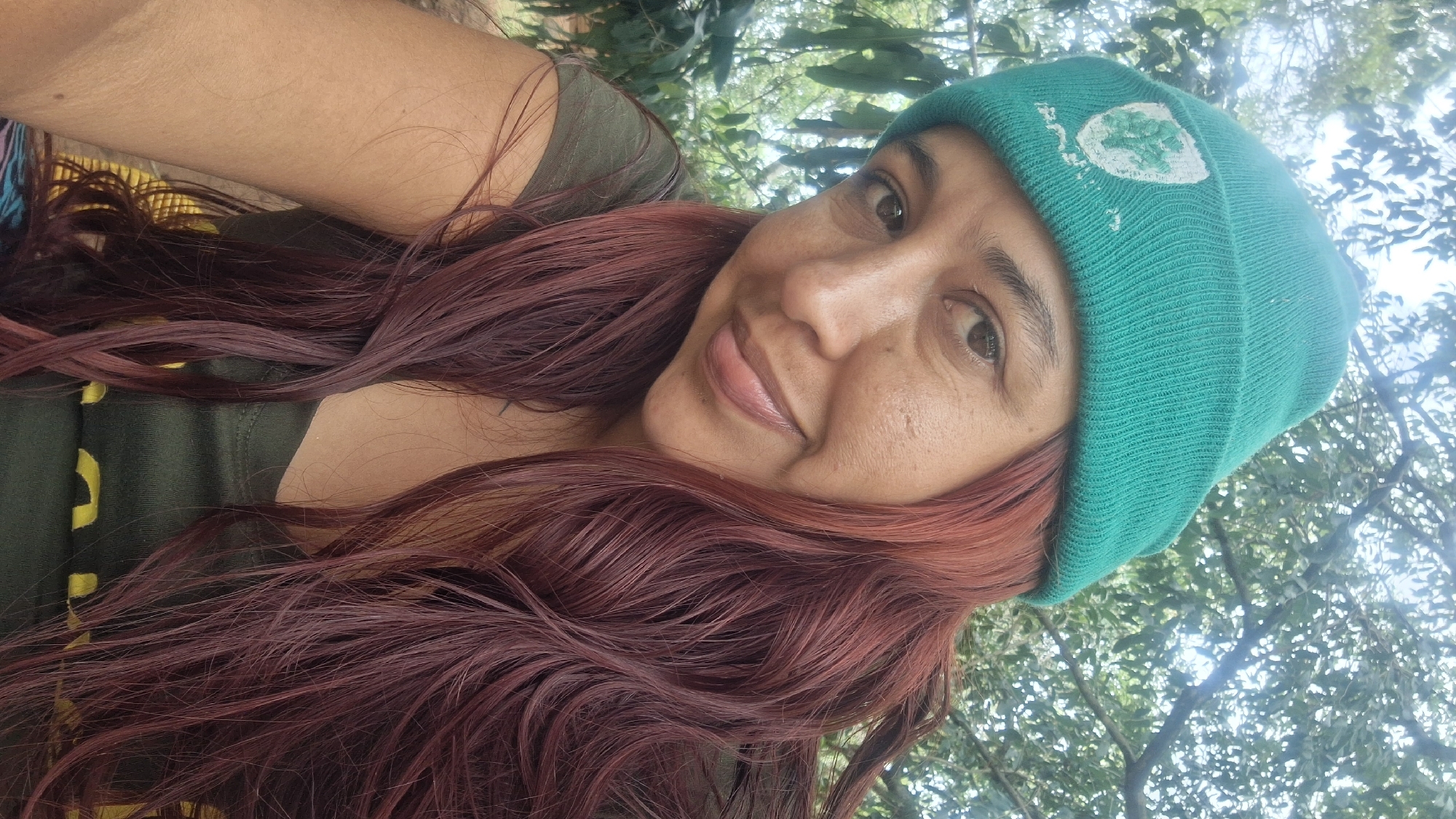 A woman is captured in a close-up selfie, oriented diagonally, set outdoors in what appears to be a natural, leafy environment in Vallecito, Venezuela. She has a medium skin tone, dark eyes, and long, wavy reddish-brown hair flowing over her shoulders. A gentle, soft smile plays on her lips as she looks towards the camera.
She is wearing a dark olive green top and a bright teal or emerald green knit beanie on her head. The beanie features a white, abstract design on the front, possibly resembling a stylized leaf or a simple shape, but it is not clearly legible as specific text. The background consists of dense green foliage and tree branches, with bright, diffused sunlight filtering through, suggesting it is daytime under a canopy of trees. The overall mood is relaxed and pleasant.