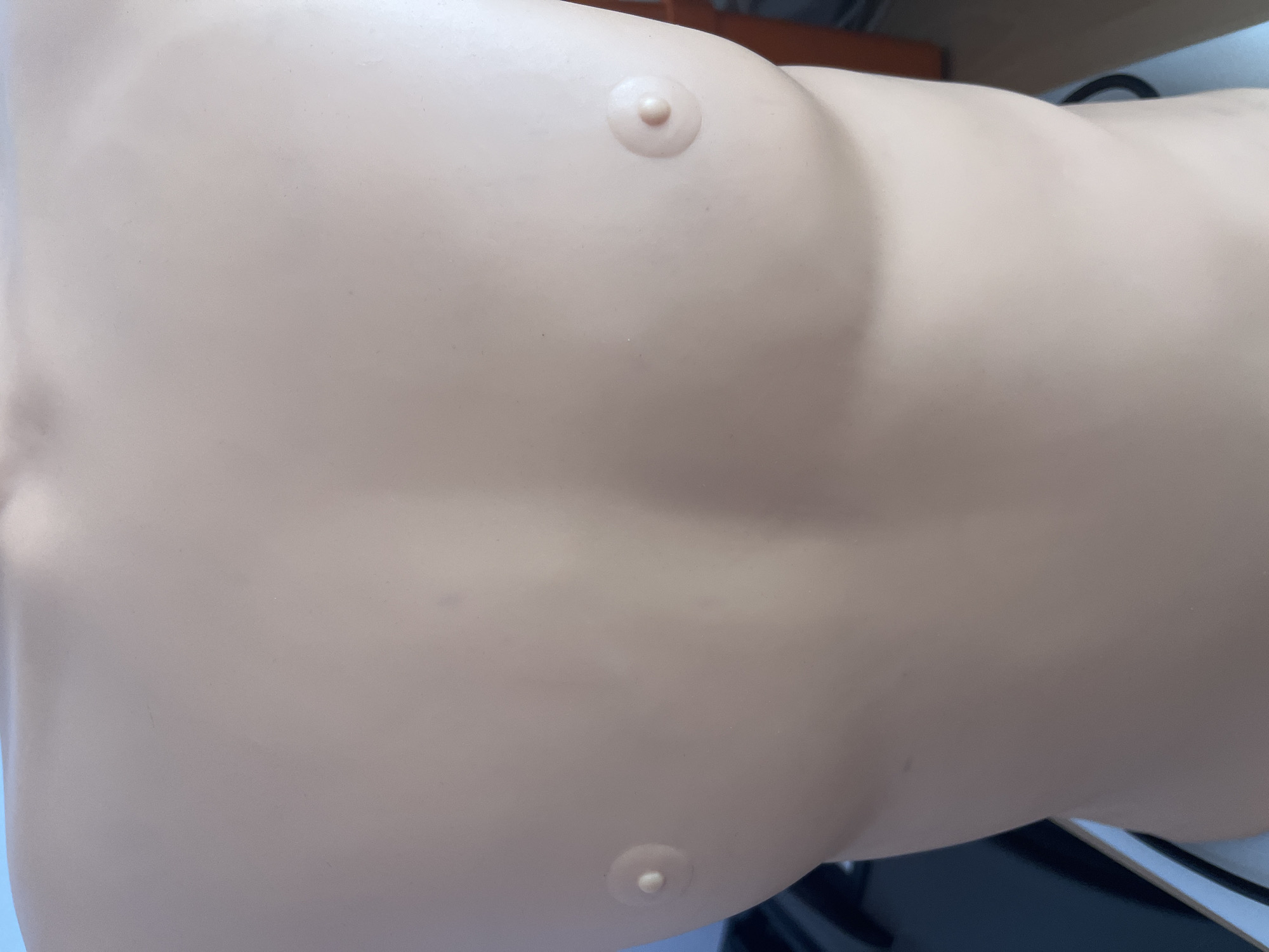 The image presents a close-up, angled view of a light-skinned, beige-colored mannequin torso, likely a medical training dummy. The smooth, uniform surface of the torso is the primary focus, devoid of any significant blemishes or markings. Two small, raised, circular protrusions are visible on the mannequin: one towards the upper right, and another towards the lower left, resembling nipples or attachment points.
The torso exhibits subtle anatomical contours, suggesting an adult human form, though it lacks specific gender-defining features other than the overall shape. The background is out of focus and indistinct, showing blurry shapes and colors that suggest an indoor setting. Darker elements are visible along the right and bottom edges of the frame, possibly furniture or equipment. The lighting is even and soft, illuminating the mannequin's surface without creating strong shadows, typical of indoor artificial light. There is no discernible activity taking place, and no text is visible anywhere in the image.