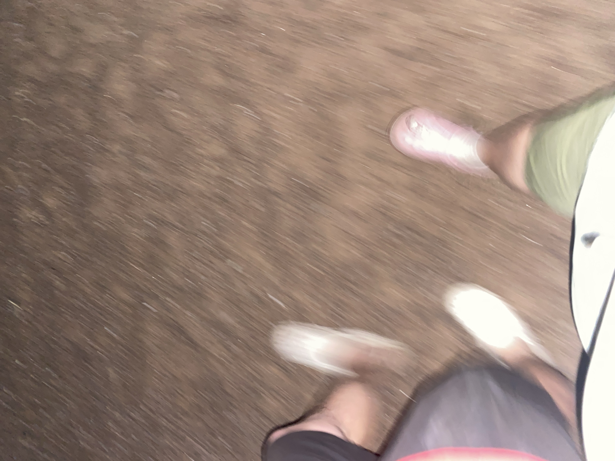 The image shows a blurred view of two people walking on a ground that appears to be dirt or sand. The left side of the image shows a person's left leg, clad in dark shorts. A blurry white shoe is visible on the right foot of this person. To the right, a second person's legs are visible, wearing light green shorts and a pink shoe. The ground is a uniform brown color with streaks indicating motion. There are no discernible buildings or urban features visible, making it impossible to identify the location as Baltiiskoe, Russia, or any specific city. The lighting suggests it is either dusk or nighttime.