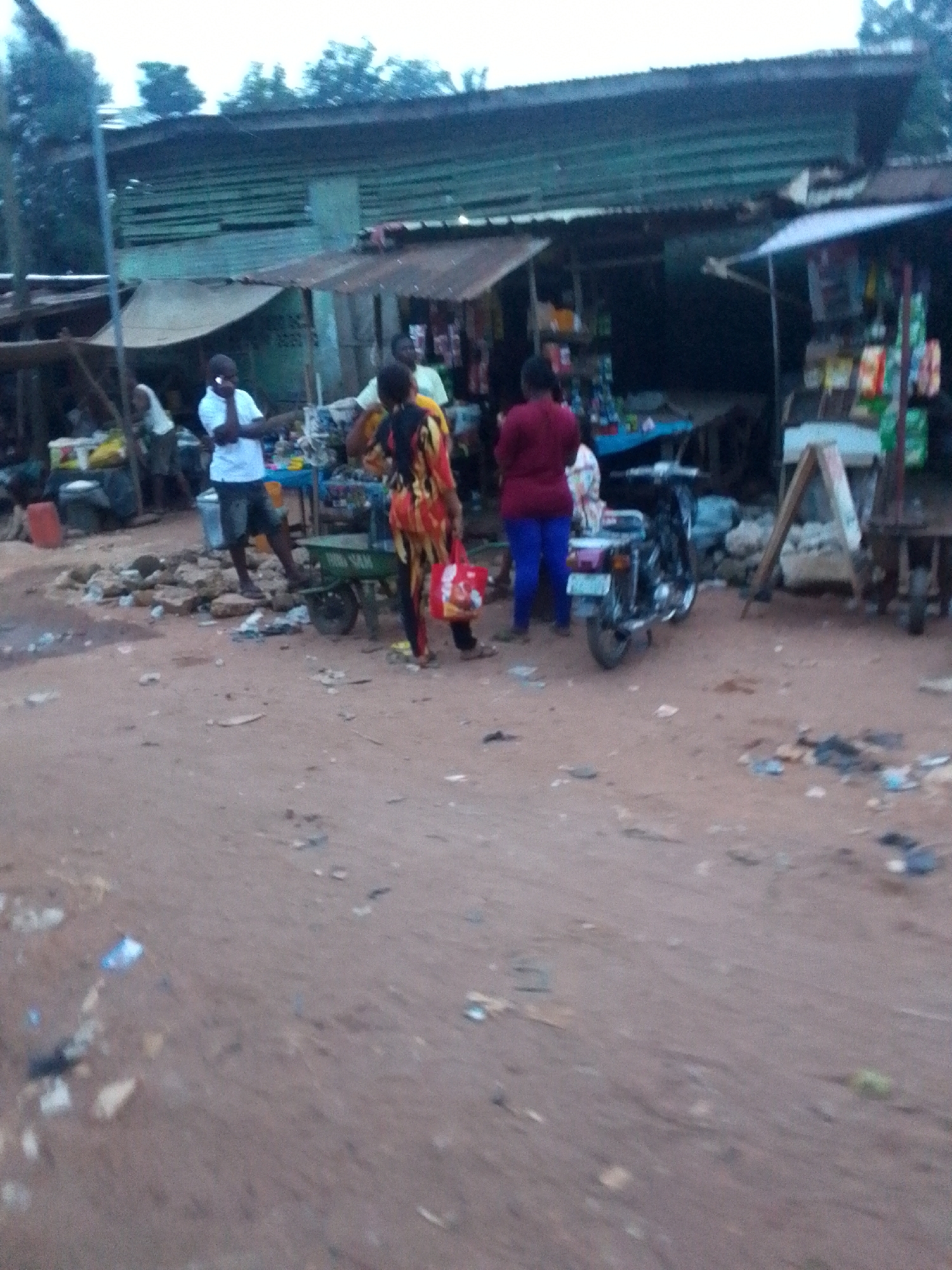 The image captures a bustling street scene, likely a market or commercial area, with an unpaved, reddish-brown dirt road in the foreground. Several people are visible, engaged in various activities.
In the center-left, a man in a white t-shirt and dark shorts stands, holding a phone to his ear. To his right, a person wearing a colorful patterned outfit (yellow, red, and black) stands next to a small green two-wheeled cart, holding a red bag. The word "RAM" is partially visible on the side of this cart. Further to the right, another person in a dark red top and blue trousers is seen near a parked motorcycle.
The background is lined with simple structures that appear to be shops or market stalls, characterized by their corrugated iron roofs. Goods are displayed, though indistinctly, on tables and shelves within these stalls. A blue table or stand is visible under one of the stalls. Rocks, debris, and scattered litter are present on the dirt ground, adding to the informal market atmosphere. Trees are visible in the distance, above the buildings.
The soft lighting suggests it might be late afternoon or early evening, or an overcast day. The scene depicts typical daily life in a semi-urban or rural setting, consistent with the provided location context of Orlu, Nigeria.