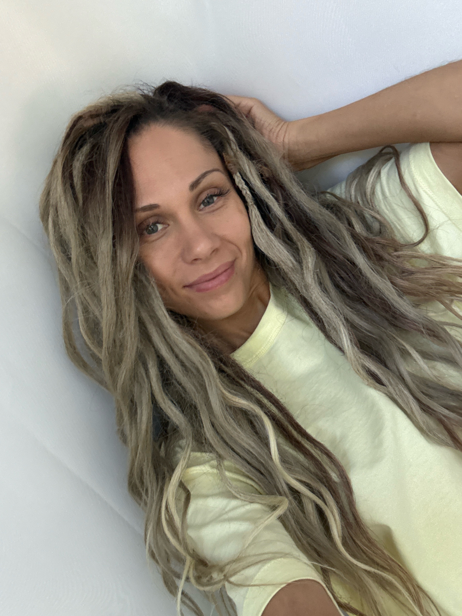 In the city of Rusanovo, Russia, a young woman with long, blonde and brown dreadlocks lies on a white surface, her head propped up by her arm. She wears a light yellow t-shirt and looks at the camera with a slight smile. Her makeup is visible, with eyeliner and mascara accentuating her blue eyes. The background is a plain white wall, with a corner visible to the left of the frame.