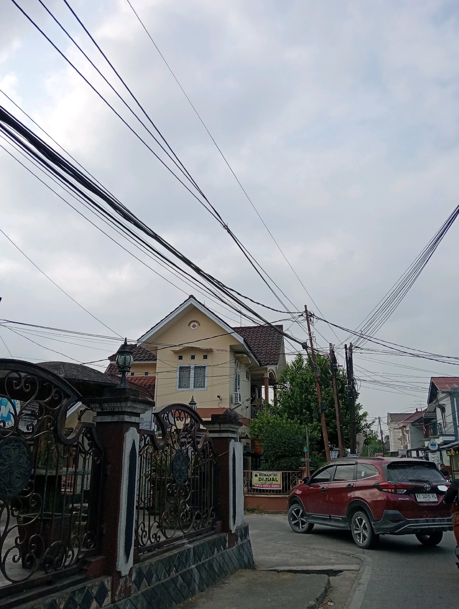 The image depicts a residential street in Samarinda, Indonesia, on an overcast day. In the foreground, a dark, ornate metal fence with stone pillars lines the left side of the frame. Behind the fence, a two-story yellow house with a red-tiled roof is visible. A red SUV, identified by its license plate "KT 1495 PL", is turning on the road in front of the house. A "RUMAH INI DIJUAL" (This House is For Sale) sign is attached to the fence near the car. The scene is characterized by numerous overhead power lines crisscrossing the sky, indicating an urban or semi-urban setting. The sky is mostly cloudy with a hint of blue. The road appears to be paved, and there are trees and other houses visible in the background.
