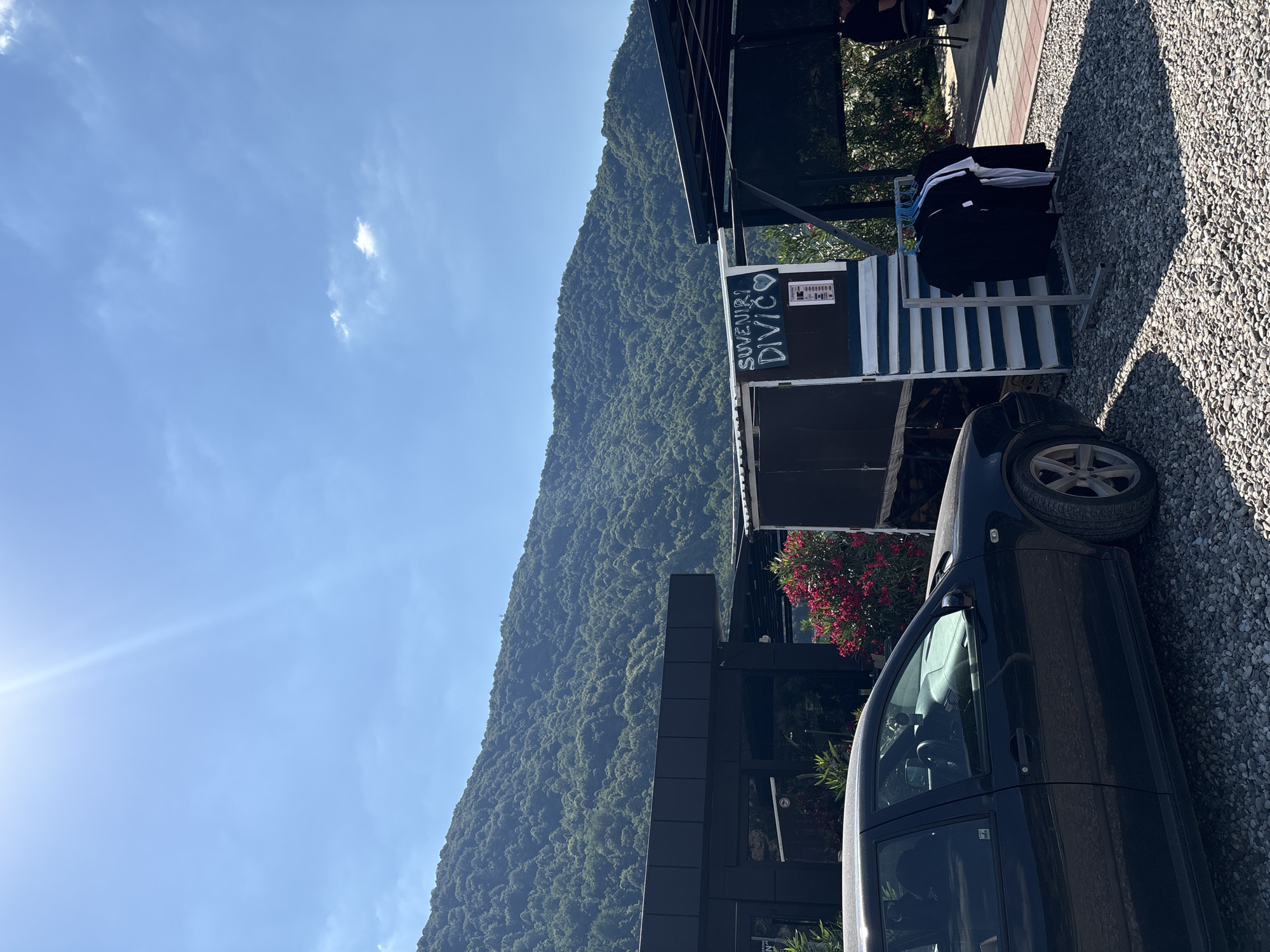 A black Volkswagen Golf is parked on a gravel lot in front of a building with a black facade. A small kiosk with blue and white stripes sells t-shirts. A sign on the kiosk reads "SUVENIRI DIVIC" with a heart symbol. In the background, a lush green mountain rises under a bright blue sky with a few scattered clouds. The scene appears to be in Divic, Bosnia and Herzegovina, given the sign.