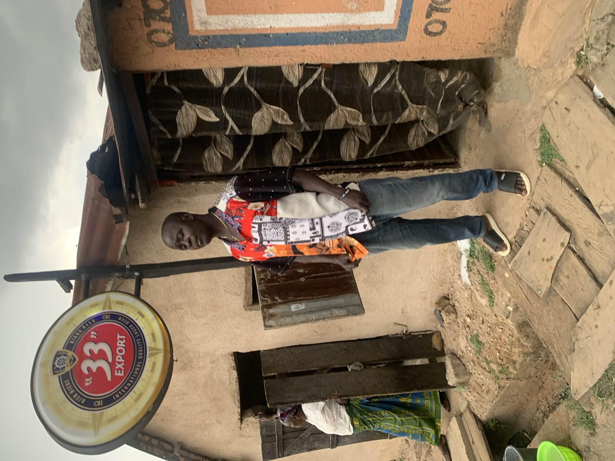The image shows a man standing in front of a building in Jos, Nigeria. The building appears to be a small, single-story structure with a weathered, light-brown exterior. A round sign advertising "33 Export" beer is mounted on the building's exterior. The man is wearing jeans and a patterned shirt. He appears to be looking at the camera. Another person wearing white is partially visible near the bottom of the image, with blue and green cloth. The ground in front of the building is uneven and appears to be a mixture of dirt and paving stones. The sky is overcast. A window is covered with decorative cloth with a leaf pattern.