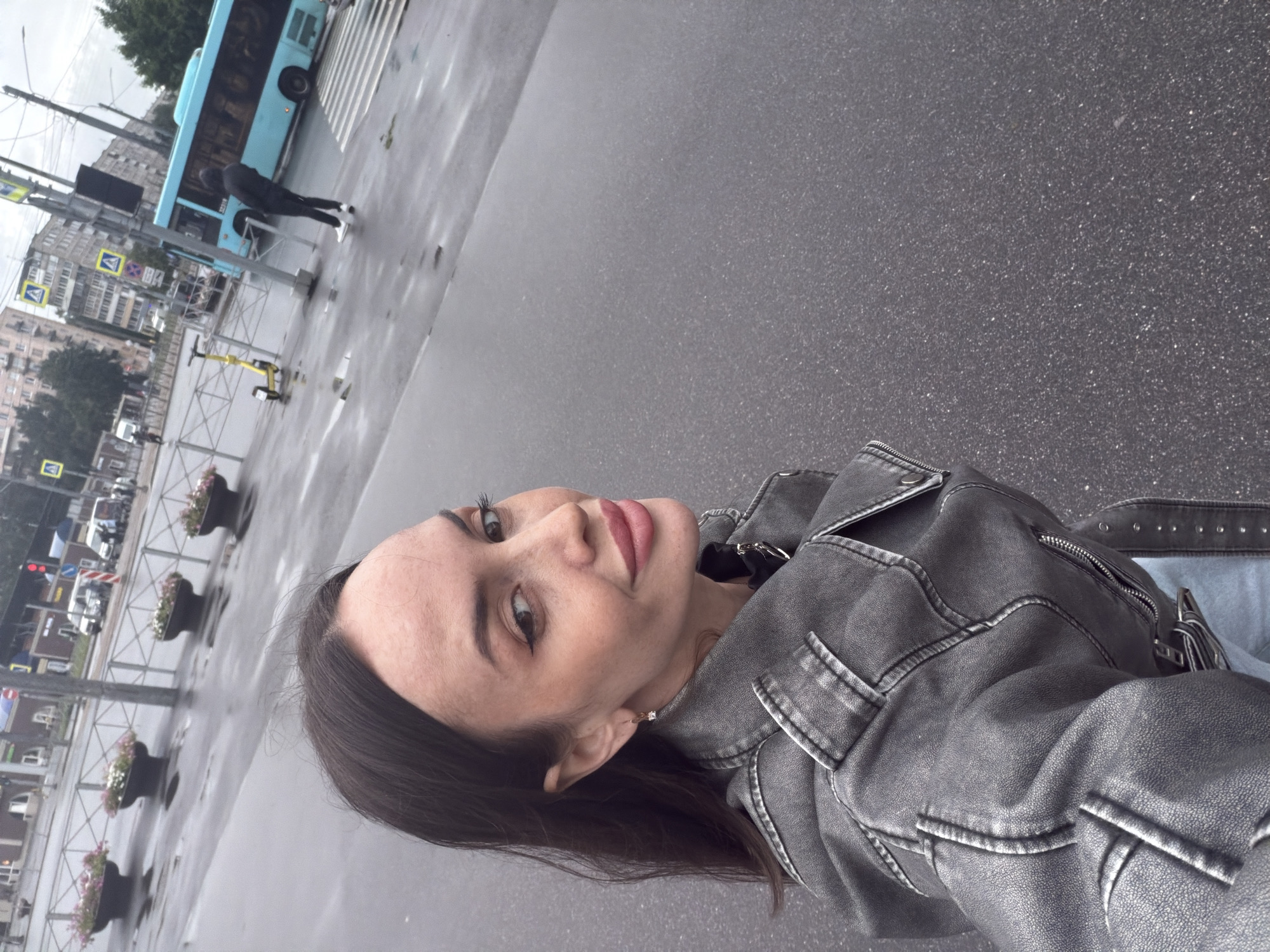 The image shows a young woman taking a selfie outdoors in what appears to be a city setting. She is wearing a grey denim jacket and has dark hair. Her expression is neutral. In the background, a blue bus is visible, as well as buildings and a crosswalk. Several traffic signs and planters with flowers are also present. The ground is wet, suggesting it has recently rained or is raining, and the sky is overcast. The lighting indicates it is daytime. The location context of Saint Petersburg, Russia, can be inferred from the visible street furniture and architectural style. There is no visible text in the image.