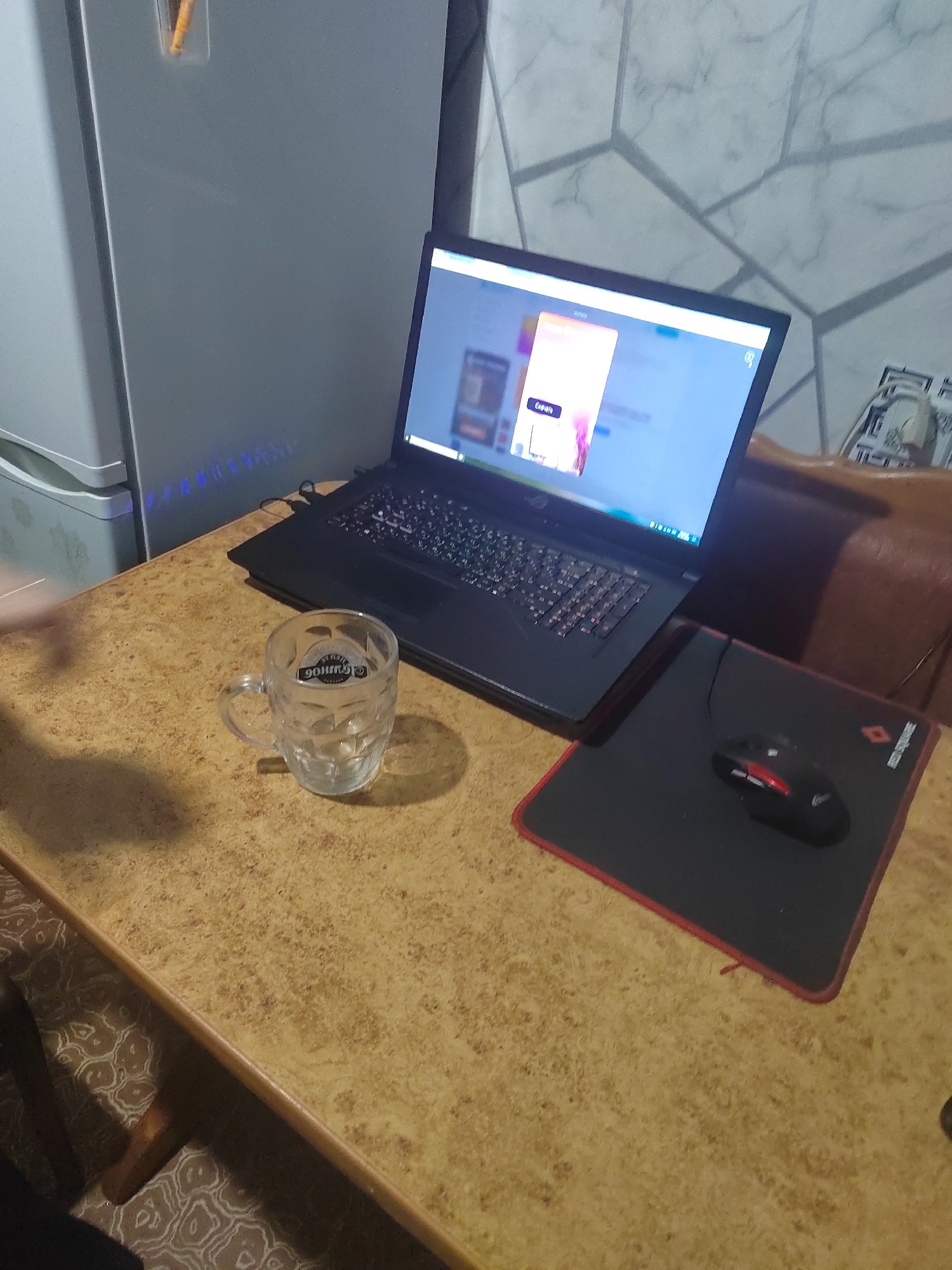 An indoor scene captured from a slightly elevated angle shows a wooden table with a speckled pattern. On the table are a laptop, a glass mug, and a mousepad with a computer mouse. The laptop, positioned at an angle, is open and displays a webpage with a pink and purple graphic. Next to the laptop, a clear glass mug with a handle sits empty. A black mousepad with red trim lies to the right of the laptop, and a black and red computer mouse is placed on it. In the background, a refrigerator is visible on the left, and a wall with a geometric pattern is on the right. The lighting suggests an indoor environment with artificial light. The scene appears to be a desk setup, possibly in a home or office. There are no people visible in the image. The image location, Slawharad, Belarus, is noted.